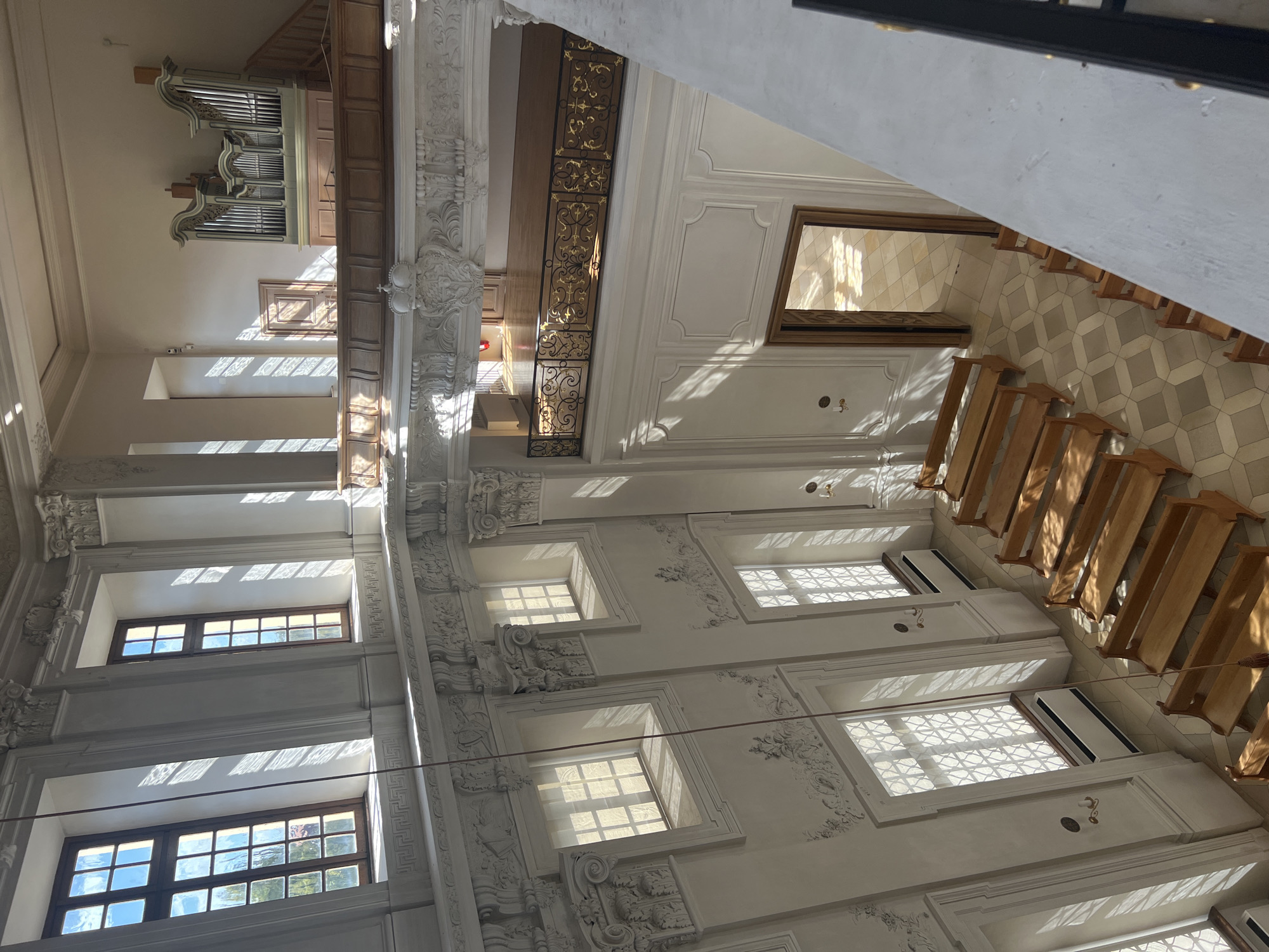 This is an interior, high-angle shot of a church or chapel in Oberschleißheim, Germany, likely part of the Schleißheim Palace complex. The space features light-colored walls with ornate white molding and decorative columns. Multiple large, rectangular windows with wooden frames and grilles are visible on the upper levels, allowing sunlight to stream in and cast patterned shadows on the walls and floor.

Below, rows of simple wooden pews are arranged on a floor with a geometric tile pattern in shades of beige and gray. The pews are empty. On one wall, a large wooden door is ajar, revealing another tiled area beyond.

In the upper right of the image, a pipe organ with light-colored pipes and a wooden casing is situated against the wall. A wooden balcony with a decorative golden railing spans across part of the upper level. The overall impression is of a well-maintained, sunlit historical interior.