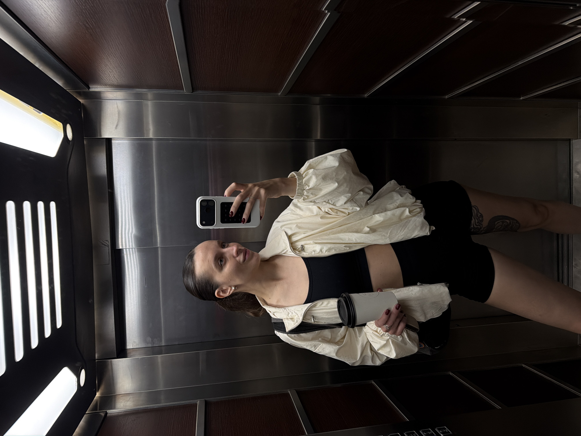 A young woman is taking a selfie in an elevator with a mirrored background, wearing a black sports bra, black shorts, and a light-colored jacket. She is holding a white cup in her left hand and a white smartphone in her right hand, showcasing manicured nails painted in dark red. A small black bag hangs on her left shoulder. Tattoos are visible on her upper thigh.
The elevator's interior consists of stainless steel and dark brown wooden panels. A lighting fixture is on the left side of the elevator. A row of elevator buttons is on the bottom right of the image.
There is no direct evidence of the location in Budva, Montenegro.