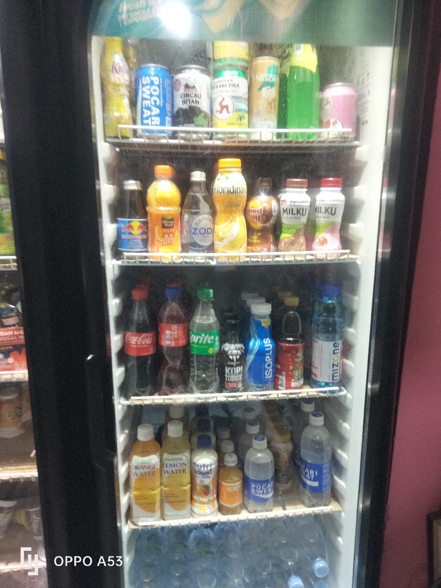 An indoor shot of a refrigerated display cabinet filled with various beverages. The cabinet is likely located in a convenience store or small shop in Karawang, Indonesia, given the presence of Indonesian-language branding on many of the products. The display features multiple shelves stocked with bottles and cans of drinks, including soft drinks like Coca-Cola and Sprite, sports drinks such as Pocari Sweat, flavored waters, milk drinks, and coffee beverages. Some of the visible brands include Pocari Sweat, Nescafe, Floridina, Milku, Coca-Cola, Sprite, and Mizone. The lighting suggests the photograph was taken in a well-lit indoor environment. The background consists of the interior of the refrigerator and a pink wall to the right. No people are visible in the image.