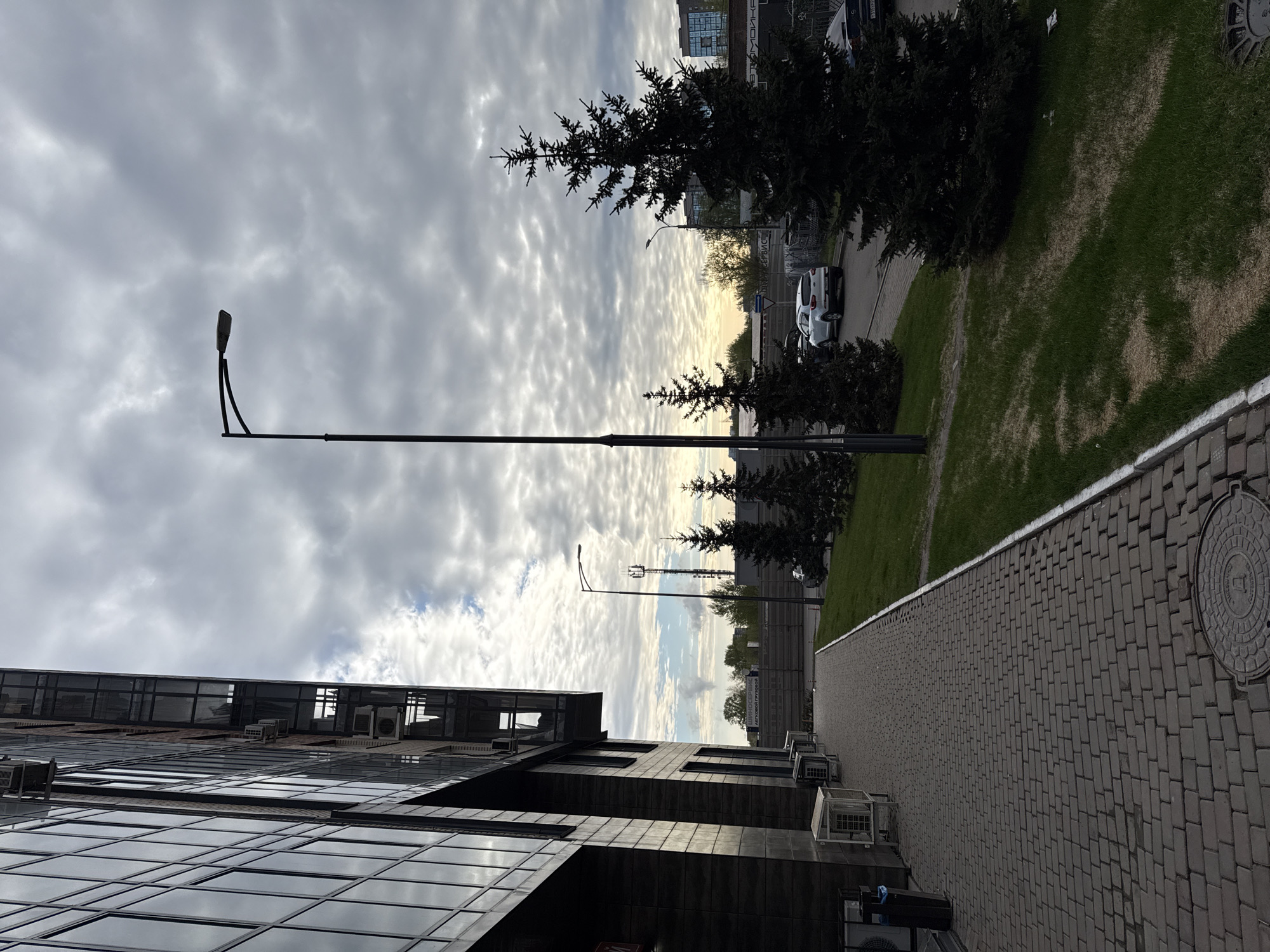 The image presents an urban scene in Krasnoyarsk, Russia. The view is taken from street level, showcasing a modern building with a dark, reflective facade on the left. The building is equipped with several air conditioning units mounted on its exterior. The foreground features a paved walkway constructed of interlocking bricks.
To the right of the walkway, there is a green grassy area with several pine trees. A row of streetlights runs along the edge of the grassy area, extending into the distance. Beyond the trees, a street with parked cars is visible.
The sky is overcast with dramatic cloud formations. The general atmosphere suggests either early morning or late afternoon due to the soft lighting. No people are visible in the frame.