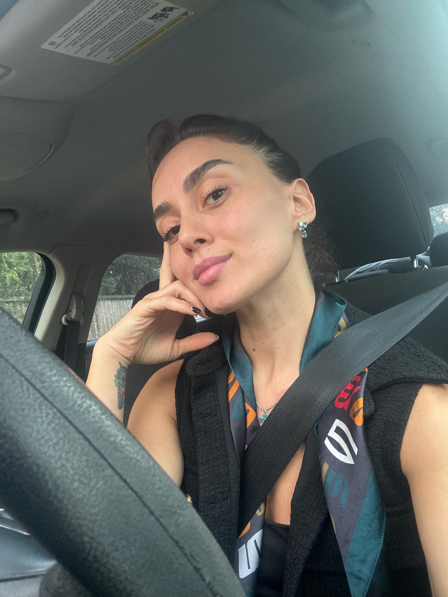A medium shot captures a woman with dark hair pulled back, seated in the driver's seat of a car. She is looking towards the camera with a slight smile, her left hand resting on her cheek. She is wearing a black vest over a dark top with a colorful scarf. A seatbelt is visible across her chest. The interior of the car, including the steering wheel and dashboard, is visible. Outside the car window, trees and a fence can be seen. The scene appears to be an outdoor setting, possibly a residential area, but there are no explicit visual cues to confirm its location in Tbilisi, Georgia.