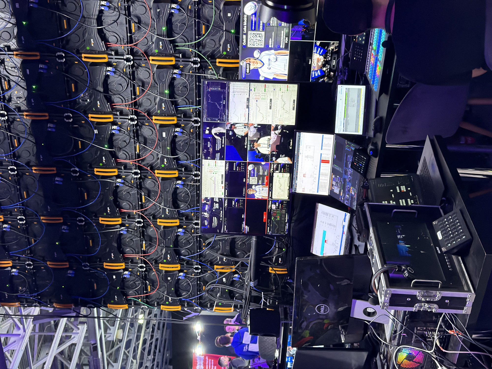 The image captures a behind-the-scenes view of a video production setup, possibly for a live event or broadcast. A large LED video wall composed of interconnected panels dominates the left side of the image. The panels feature visible cabling and connections.
To the right of the video wall, a control area is set up with multiple computer monitors displaying various data, including what appears to be charts, graphs, and video feeds related to the event's content. Some monitors feature images of individuals and brands, including "CoinW Luka Modrić."
Electronic equipment like mixers and controllers are visible, along with a Dell laptop. People are present at the bottom of the image, partially obscured, facing toward a red banner.
The text on the screens include the words "СКАНИРУЙ," "Cifra," "PGM," "CoinW," "45:07," "Luka Modrić," and "Abyen."
The image depicts a technical environment indicative of media production, likely in a city such as Khimki, Russia. The image does not depict a specific location.