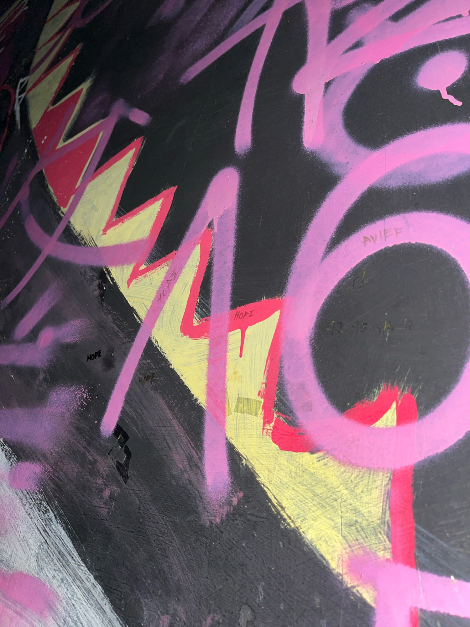 The image shows a close-up of graffiti on a dark, textured surface, possibly a wall. The graffiti features large, bold strokes of pink and purple spray paint, with some areas appearing to be layered or overlapping. Yellow and red elements are also present, creating sharp, angular shapes that contrast with the smoother curves of the pink spray paint. Faintly visible text appears in several areas, including the word "HOPE" written in black, and other illegible markings. The overall impression is one of vibrant, chaotic street art. The location in Surakarta, Indonesia is not evident from the image itself.