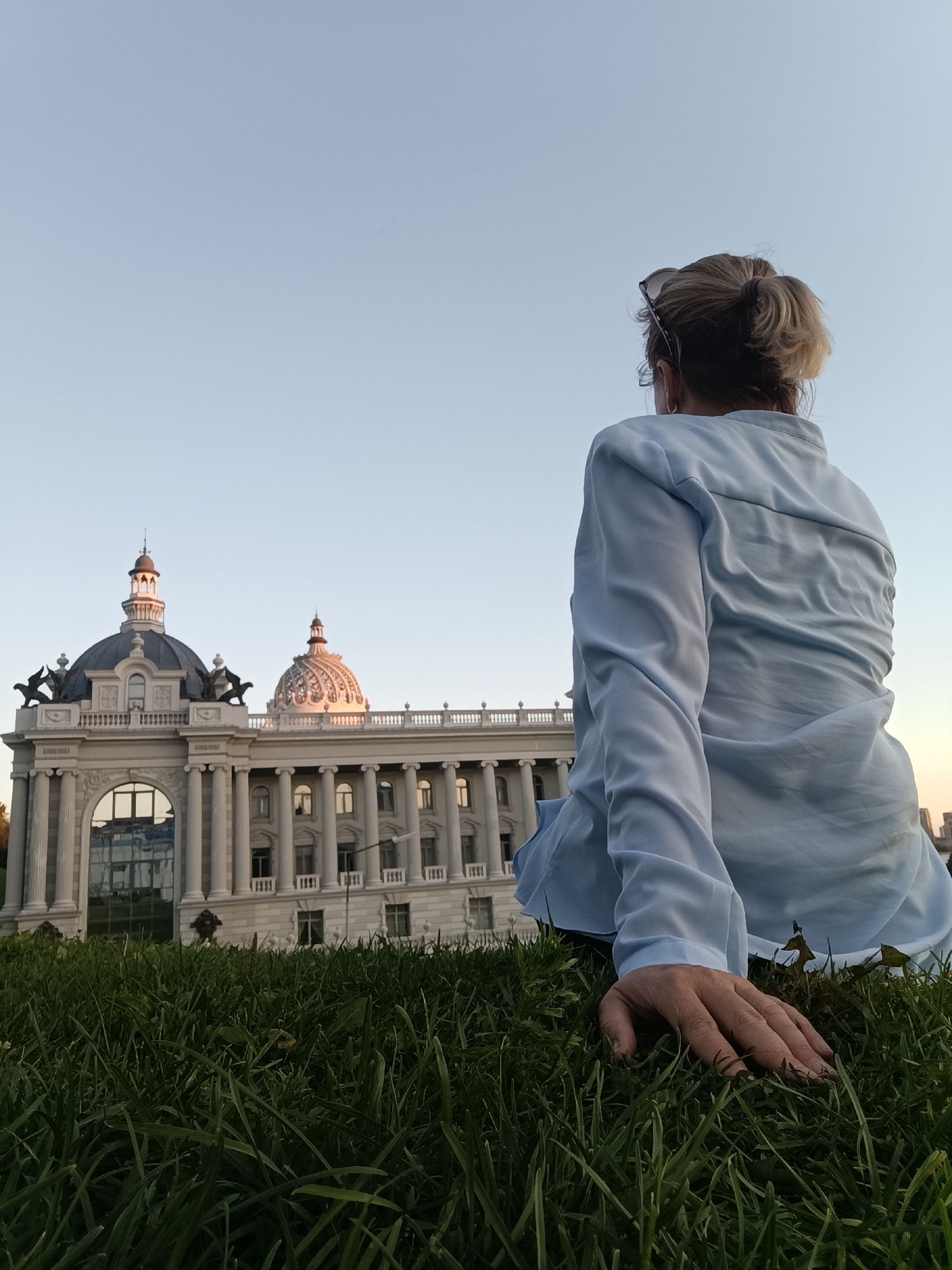 A woman with blonde hair in a bun, wearing sunglasses on her head and a light blue button-up shirt, sits on a grassy lawn. She is viewed from behind, facing a building with classical architecture. The building features multiple columns, balconies, and two distinct domes, one dark and one lighter with intricate detailing. Statues of winged lions adorn the roofline of the building. The sky is clear and pale blue, transitioning to a subtle orange hue at the horizon on the right side of the image. The foreground consists of dense green grass. The image appears to be taken at a low angle, emphasizing the building and the sky. This is the National Library of the Republic of Tatarstan in Kazan, Russia.