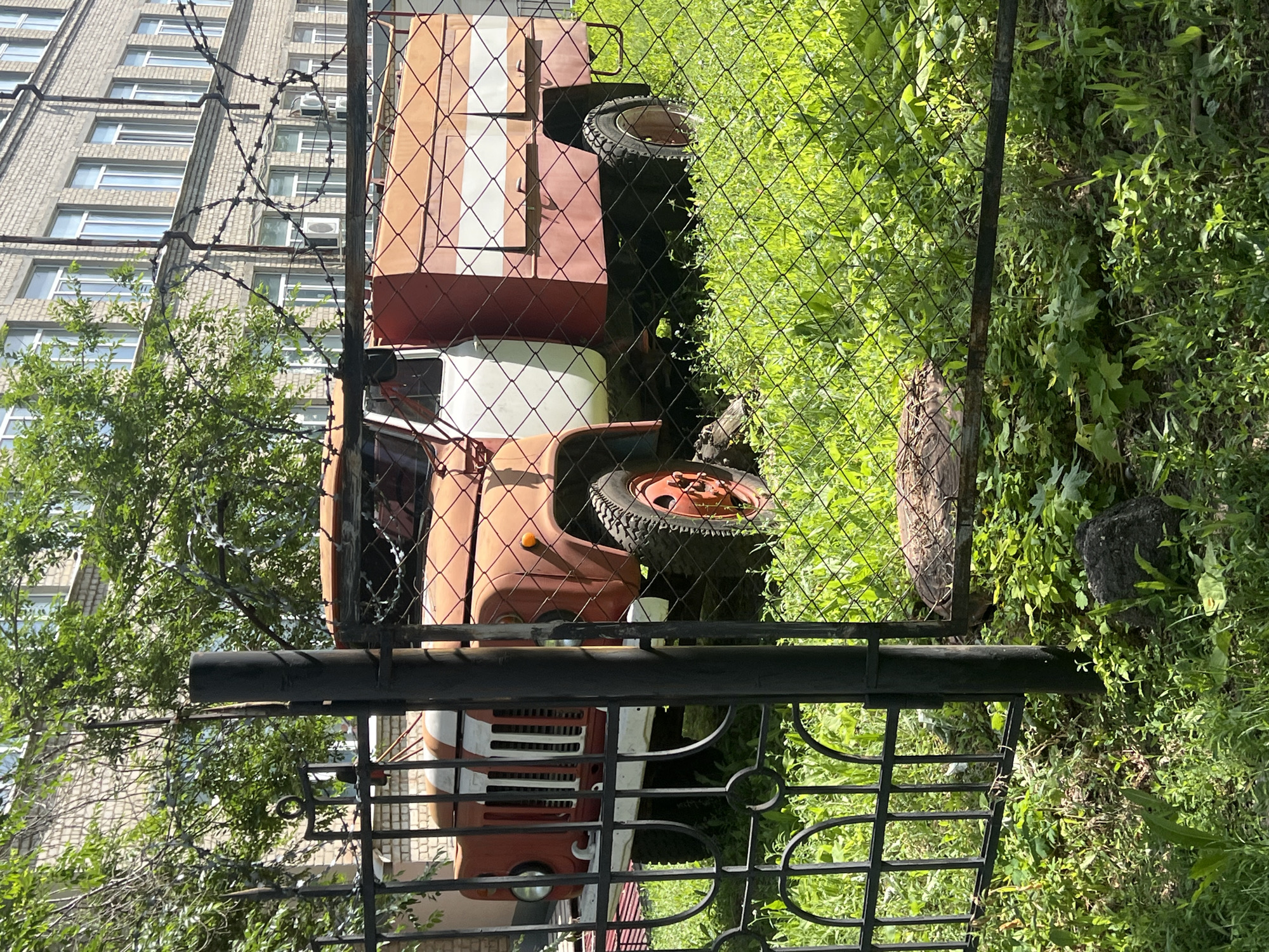 An abandoned, rusty, orange and white truck with a utility bed is parked on a grassy area behind a black metal fence. The truck has a prominent grill and a visible tire. In the background, a brick building with multiple windows and trees with green leaves are visible. Barbed wire is strung along the top of the fence. The ground is covered in green grass and some rocks.