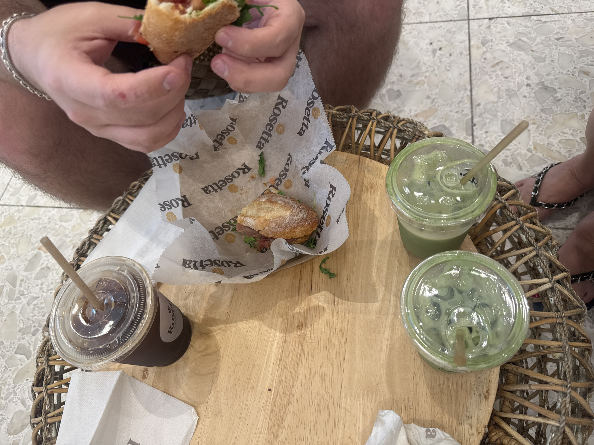 The image shows a top-down view of a group of people eating and drinking at a table. A person is holding a sandwich with both hands. Another sandwich sits in a paper wrapper labeled "Rosetta." There are two green drinks in plastic cups with lids and straws, and one brown drink in a similar cup. The table is made of light-colored wood and is placed on a wicker base. The background shows a tiled floor. The location is likely a restaurant in Aventura, United States of America, based on the "Rosetta" branding, which is a known restaurant in Aventura.