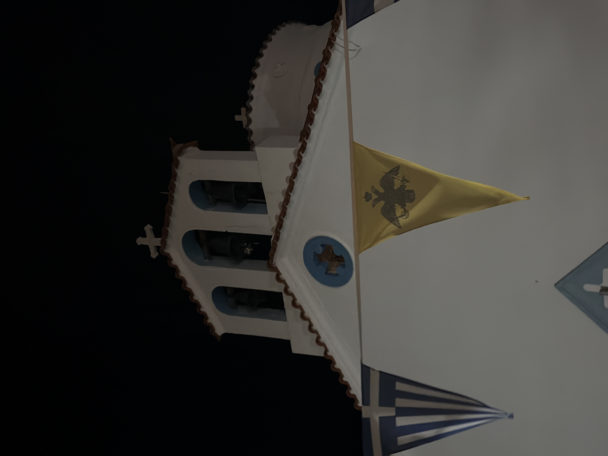 The image shows a close-up of a white church building at night, with a dark sky. The church has a bell tower with three arched openings, each containing a bell. A cross is visible at the apex of the bell tower. The roof of the church is tiled with reddish-brown tiles. Below the bell tower, there is a triangular section of the facade with a blue circular emblem featuring a cross. Two triangular flags are hanging in front of the church. The flag on the left is the Greek flag, with blue and white stripes. The flag on the right is yellow and features a black double-headed eagle emblem. The image does not provide any specific location details within the city of Βιγκλάφια, Greece, other than it is a church in that city.