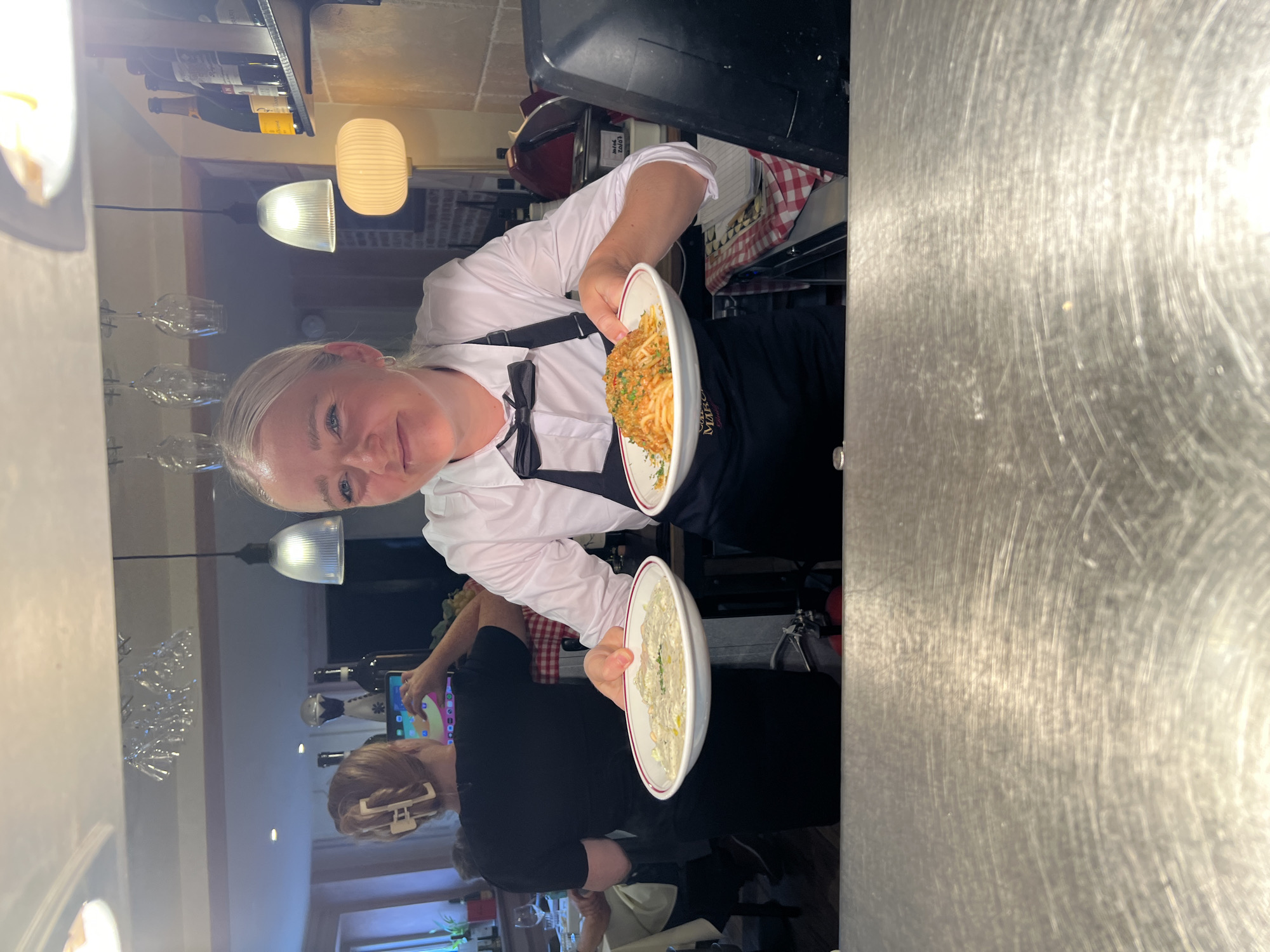 A blonde waitress, wearing a white shirt, black bow tie, and black apron, holds two plates of food in a restaurant. The plate on her left contains a pale, creamy dish with green herbs sprinkled on top. The plate on her right holds a portion of spaghetti with a reddish sauce and breadcrumbs. In the background, another person in a black shirt and a white hair clip is visible using a tablet. The restaurant environment includes overhead lights, wine glasses hanging from a rack, shelves stocked with bottles, and a stainless steel counter in the foreground. The overall scene depicts a restaurant kitchen or service area, likely in Guildford, United Kingdom, given the context.