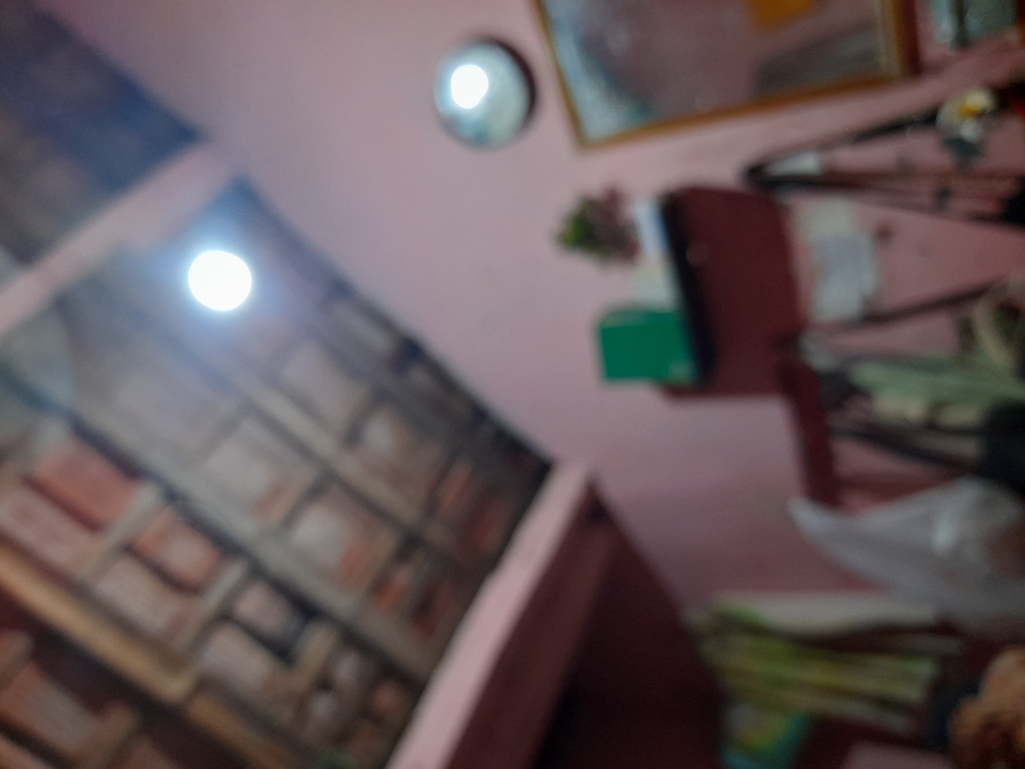 The image depicts an interior scene, likely a room, with a prominent bright light source in the upper left quadrant. The lighting suggests it might be nighttime or a dimly lit interior space. The overall perspective is looking upwards towards the ceiling and upper walls.
In the foreground and left side of the image, there appears to be a wall covered in a textured pattern that resembles bricks or tiles, laid in a herringbone pattern. This wall is angled and extends towards the viewer.
On the upper right side, the scene is more cluttered, suggesting a shelf or a desk area. Several objects are visible but blurred due to the image's focus. These include what seems to be a framed picture on the pink-colored wall, a small green object that could be a book or folder, and a dark red object that might be another book or a piece of furniture. There are also stacks of items, possibly papers or books, and a white plastic bag.
The overall setting appears to be a domestic or storage space. The lighting is a key element, with the bright light drawing attention and creating strong highlights and shadows, contributing to the blurred effect of the surrounding objects. There is no visible text within the image. The context of Pulokulon, Indonesia, might suggest a warm climate, but no specific weather cues are discernible from the image itself. The emotion conveyed is neutral, primarily due to the lack of clear subjects and the abstract nature of the blurred details.