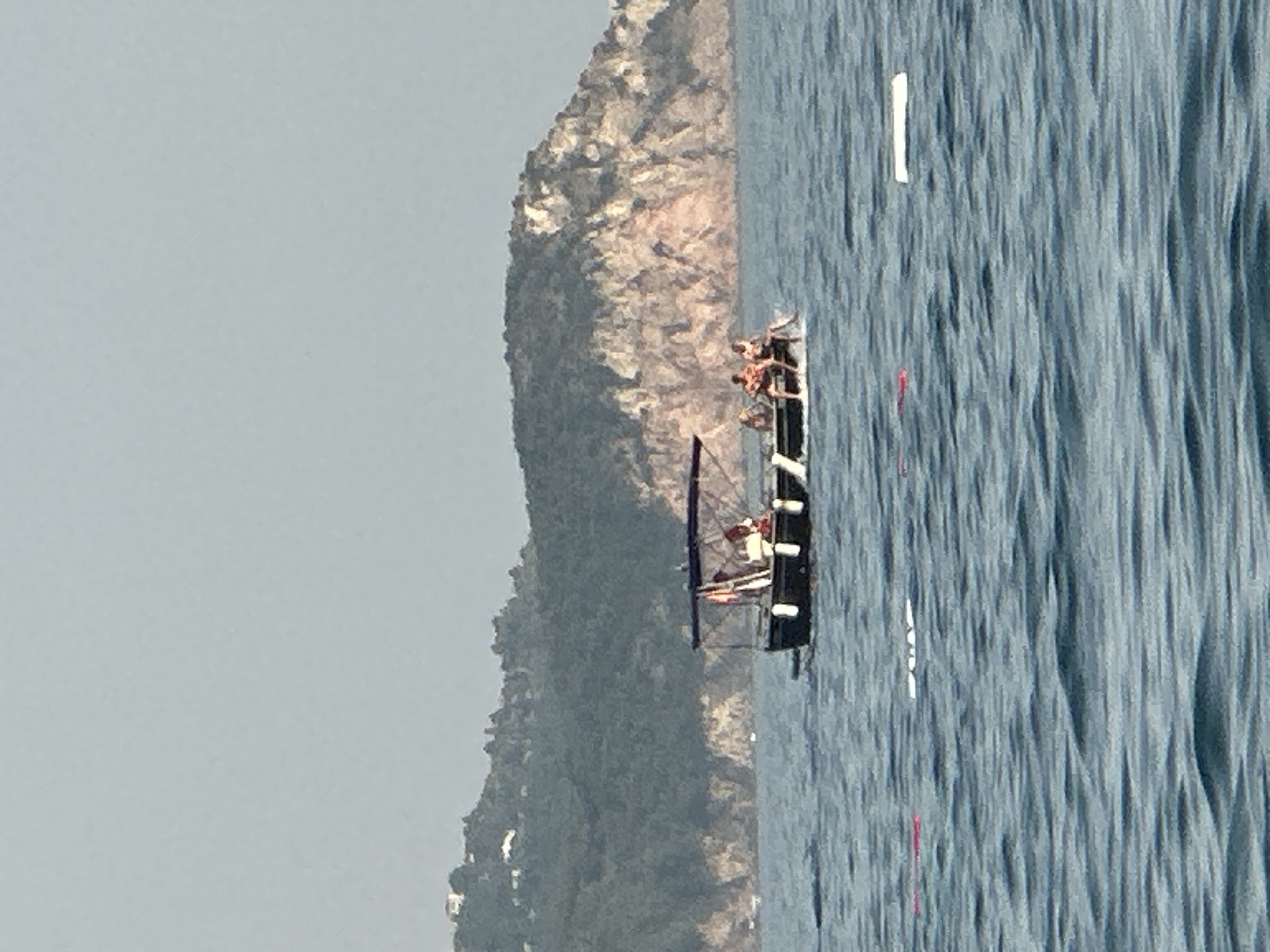 An eye-level medium shot captures a boat navigating the choppy, blue waters off the coast of Boreti, Montenegro. The boat, a dark-hulled vessel with a white trim and a canopy, carries four individuals. Two people are seated towards the rear of the boat, and two are sitting on the edge of the boat at the front. The background features a verdant, mountainous landscape dotted with houses. The sky is a pale, hazy blue. The water is disturbed by moderate waves, and in the foreground, several buoys or markers are visible, some red and some white. The overall scene depicts a leisurely day on the water with a backdrop of coastal Montenegro.