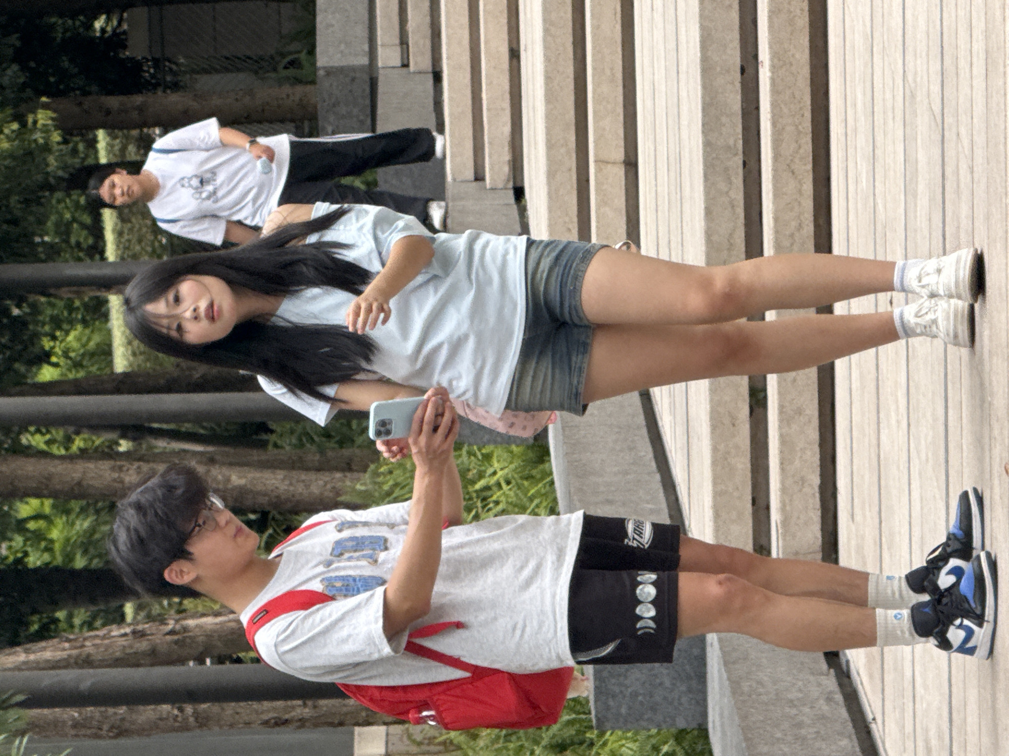 In Yuzhong District, China, a young Asian male with glasses and dark hair, wearing a gray t-shirt, black shorts with a moon phase graphic, and blue and white sneakers, stands on a wooden walkway. He is holding a light blue smartphone and looking down at it. To his right, a young Asian female with long dark hair, wearing a light blue off-the-shoulder top and denim shorts, looks forward. Behind them, another young Asian female in a white t-shirt with a graphic and black pants with white stripes walks up a set of concrete stairs. The background features trees and foliage, suggesting an outdoor urban setting.