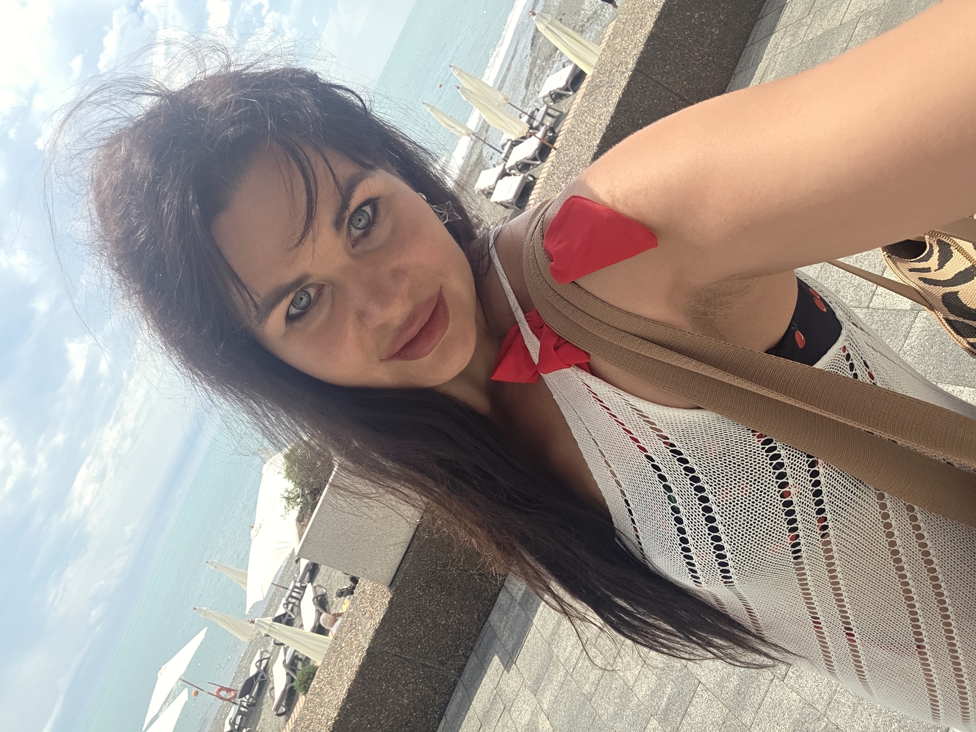 A woman with dark hair and blue eyes takes a selfie on a sunny day at a beach resort in Sochi, Russia. She is wearing a white, crocheted cover-up over a black bikini with a red fruit pattern. A red bow is attached to the strap of her cover-up, and a red heart-shaped object is pinned to her shoulder. She is carrying a brown bag with a leopard print accent. In the background, lounge chairs and white umbrellas are set up on a pebbled beach, and the turquoise sea stretches to the horizon under a cloudy sky. The foreground shows a paved walkway.
