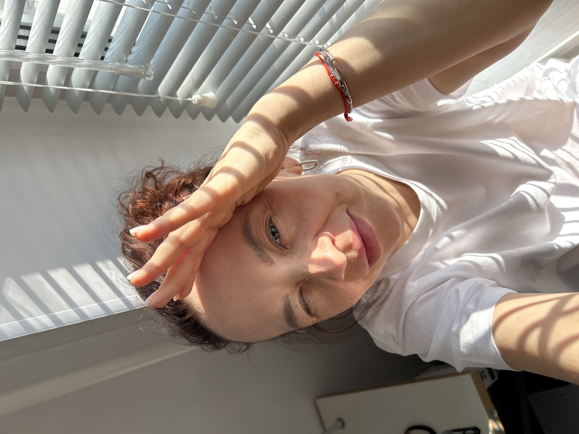 A light-skinned woman with curly reddish-brown hair is indoors next to a window with closed white blinds. She is wearing a white t-shirt and has a bracelet with red and silver components on her raised right wrist. Her right hand is shielding her face from the sunlight, and her head is tilted toward the right. Her lips are slightly parted, and she has one eye closed. The location is likely Chaikovsky, Russia.