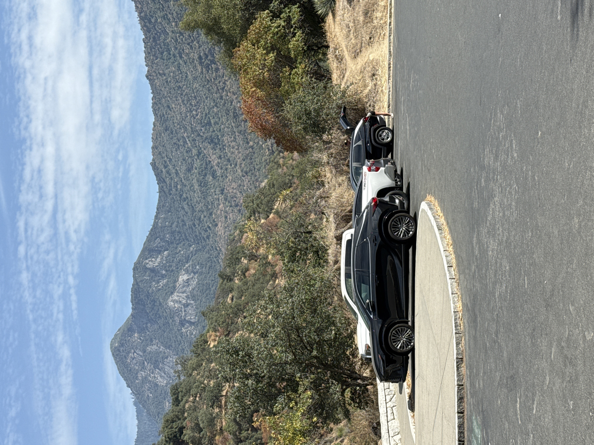 In Three Rivers, California, United States, a black Lexus SUV is parked on asphalt next to a white truck and a black Toyota SUV in a parking lot. The Lexus is in the foreground, facing right, with its wheels turned. The white truck is parked behind the Lexus, and the black Toyota SUV is parked behind the truck. The Toyota's trunk is open, and a person in a white shirt and dark shorts is standing next to it, seemingly loading or unloading items. The parking lot is situated on a gentle slope with a stone retaining wall lining the edge of the parking area. Beyond the parking lot, the terrain rises into a hillside covered with dry grasses and scattered trees. Further in the background, a range of mountains dominates the landscape, with one prominent peak in the center. The sky above is blue with scattered white clouds. The overall environment suggests a park or a scenic overlook in a mountainous region.