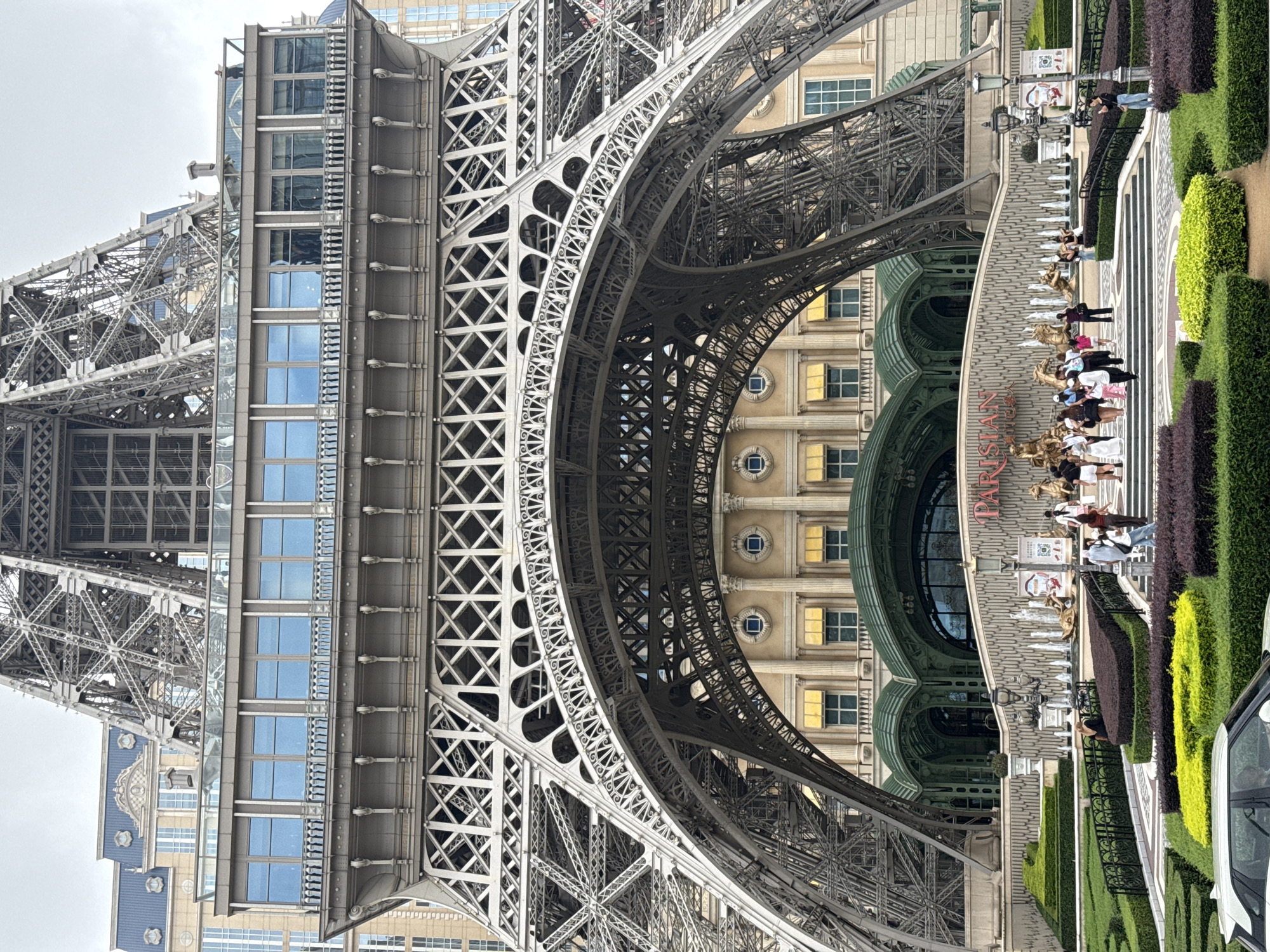 The image shows a partial view of the Parisian Macao, including the facade of the hotel and the Eiffel Tower replica. People are walking along a path with manicured hedges and fountains leading towards the hotel entrance. The sky appears overcast. A car is visible on the right side of the image. The tower structure is steel lattice.