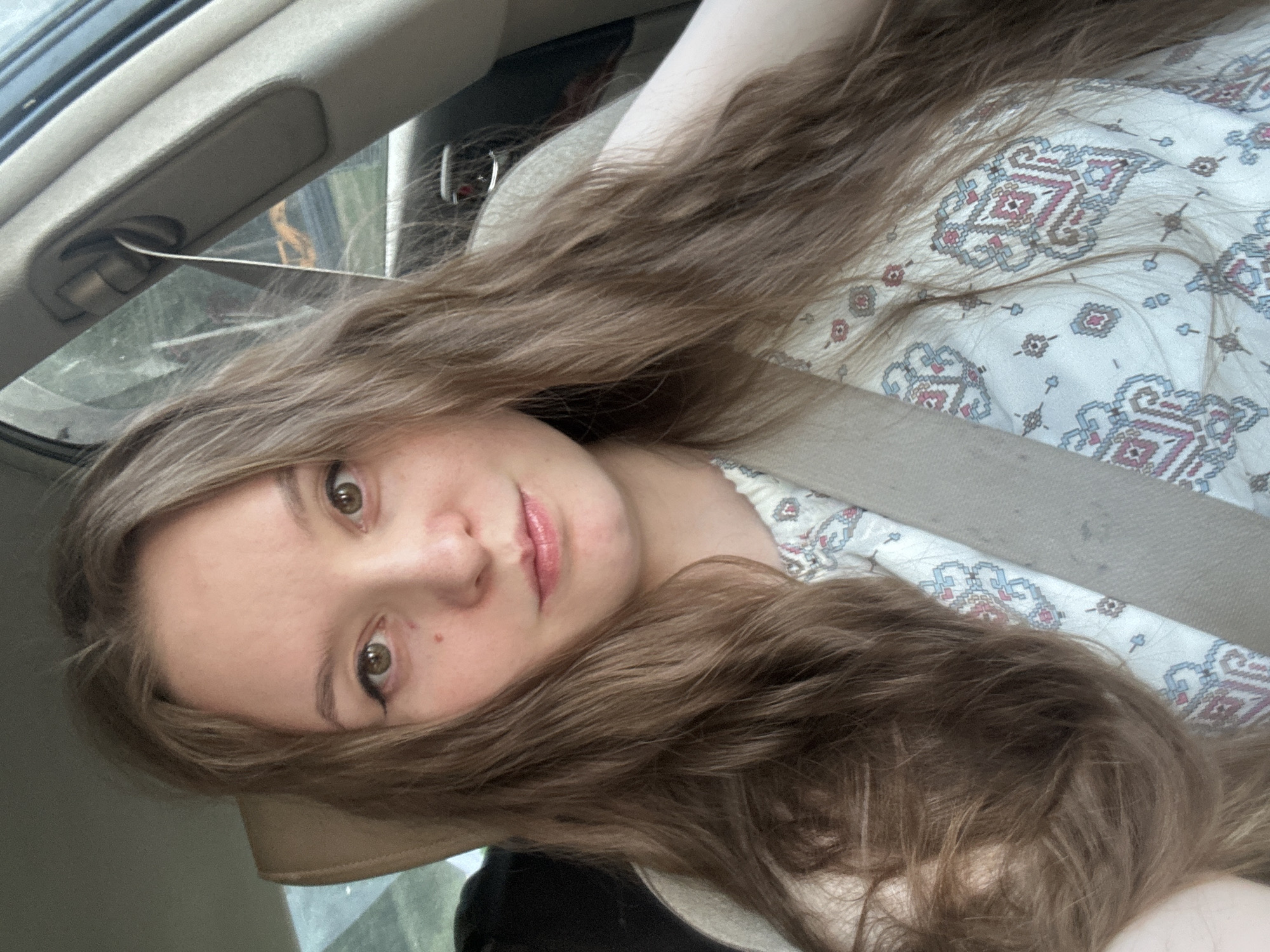 A young woman is in the passenger seat of a car, wearing a seatbelt. She has long, wavy brown hair and is looking directly at the camera. She is wearing a light-colored patterned shirt. The interior of the car is visible, including the headrest and part of the dashboard. The location is not discernible from the image.