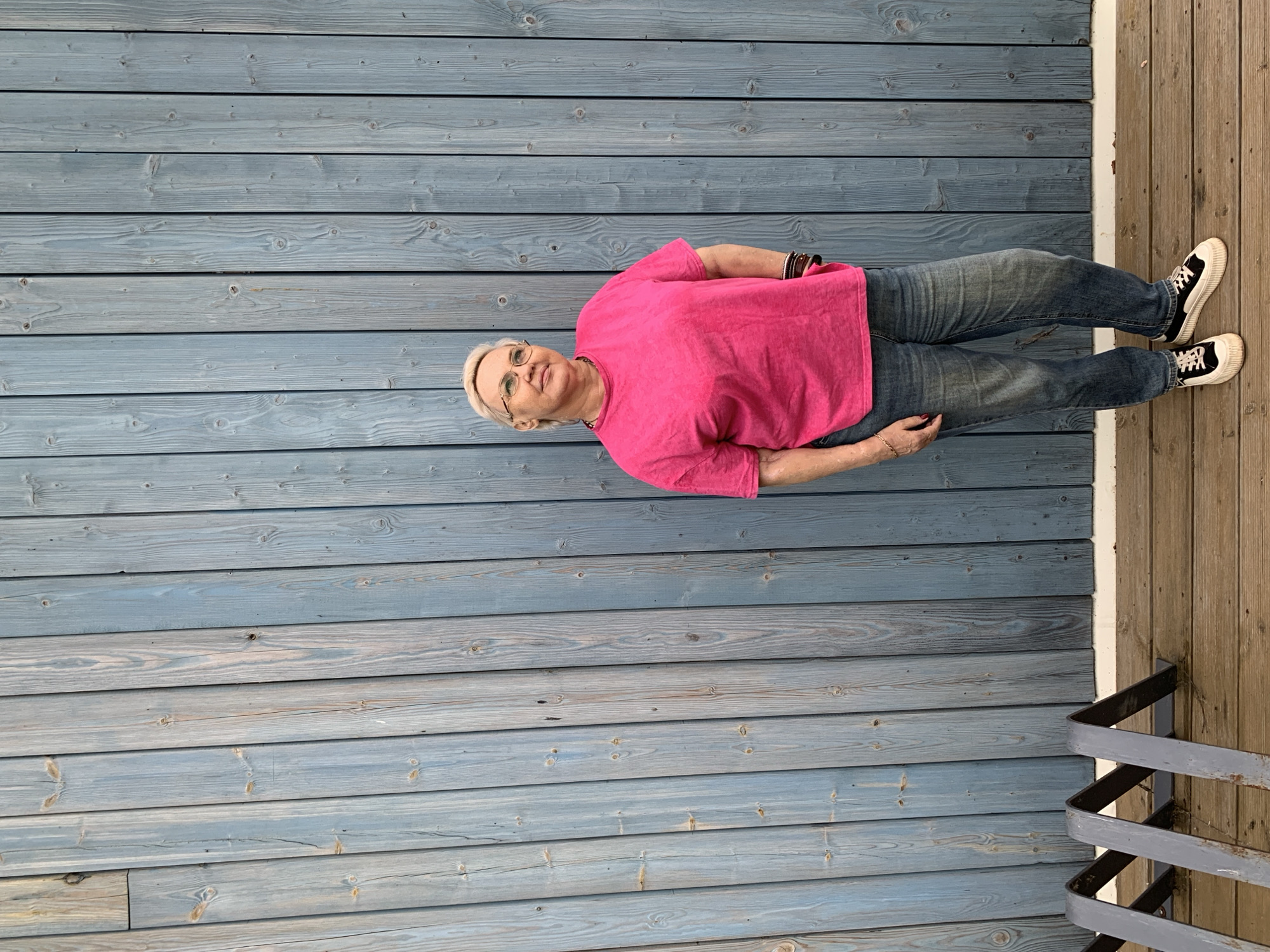 The image depicts a woman standing outdoors in Chaikovsky, Russia. She is a middle-aged to senior woman with short, light blonde hair and round, thin-framed glasses. Her gaze is directed upwards and to her left (viewer's right), with a relaxed expression. She wears a bright fuchsia pink, short-sleeved t-shirt, loose-fitting blue denim jeans that are slightly faded and cuffed at the ankles, and black and white platform sneakers with thick white soles. Accessories include a dark, multi-strand necklace, multiple dark bracelets on her left wrist, and a thin gold chain on her right wrist. Her left hand is partially in her jeans pocket, while her right arm hangs loosely, with her hand also near or in a pocket.
The background consists of a vertical wall made of weathered, muted blue-grey wooden planks. The planks exhibit varied wood grain and knots, with some appearing more faded or showing natural wood tones. Below the wall is a wooden deck floor of warm brown planks, separated by a thin white trim board. In the bottom-left foreground, a grey metal structure, consistent with a bicycle rack, is visible, forming angular, zigzag patterns.
