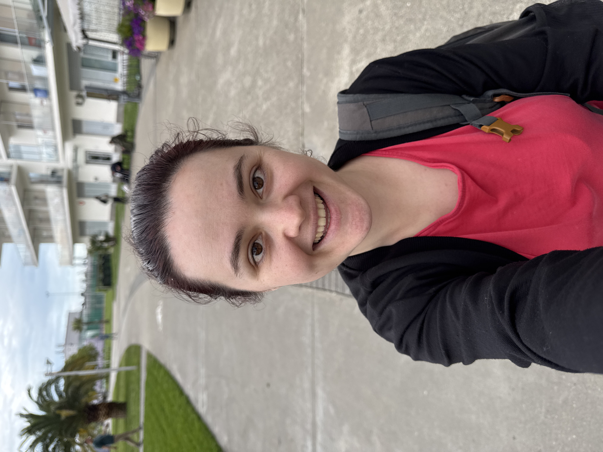 A woman wearing a red shirt and black cardigan is taking a selfie. She has dark hair pulled back and is smiling. She is wearing a backpack. In the background, there are apartment buildings, palm trees, and a grey paved surface with green grass areas. The scene is likely outdoors during the daytime. Based on the architecture and vegetation, the location is possibly in Sochi, Russia.