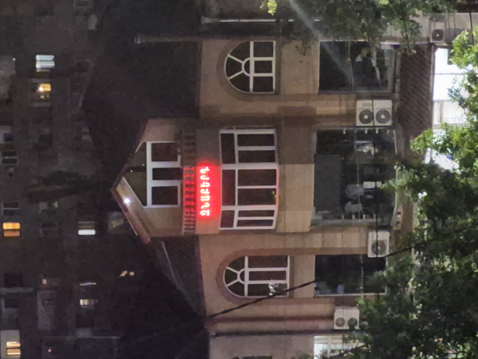 This is a nighttime, exterior view of a multi-story building located in an urban area of Yerevan, Armenia.
The central subject is a light-brown facade building, illuminated by artificial lights. Its upper section features a central gabled roof element with three tall, narrow rectangular windows below it. Directly beneath these windows, on a balcony-like projection, a red neon sign displays the Armenian text "ՁԱԽԵՐԻ" (Dzakheri), which translates to "of the lefts" or "on the left side" (plural genitive). Below the neon sign, there is a prominent bay window structure composed of multiple rectangular panes. Flanking this bay window, to the left and right on the same level, are large arched windows with white frames and radial divisions in their upper curved sections. The ground floor appears to consist of large glass storefronts that reflect ambient light and indistinct shapes from within.
Two white exterior air conditioning units are mounted on the building's facade below the arched window on the right side. Another similar unit is partially visible on the left side, below a darker, possibly reflective, window. The lower foreground is partially obscured by dark green foliage, suggesting trees or shrubs. In the background, behind the main building, several indistinct, darker buildings, consistent with apartment blocks, exhibit multiple illuminated rectangular windows. No specific individuals or actions are discernible within the image. The overall scene is characterized by dim ambient light with prominent bright spots from the neon sign and internal building lights.