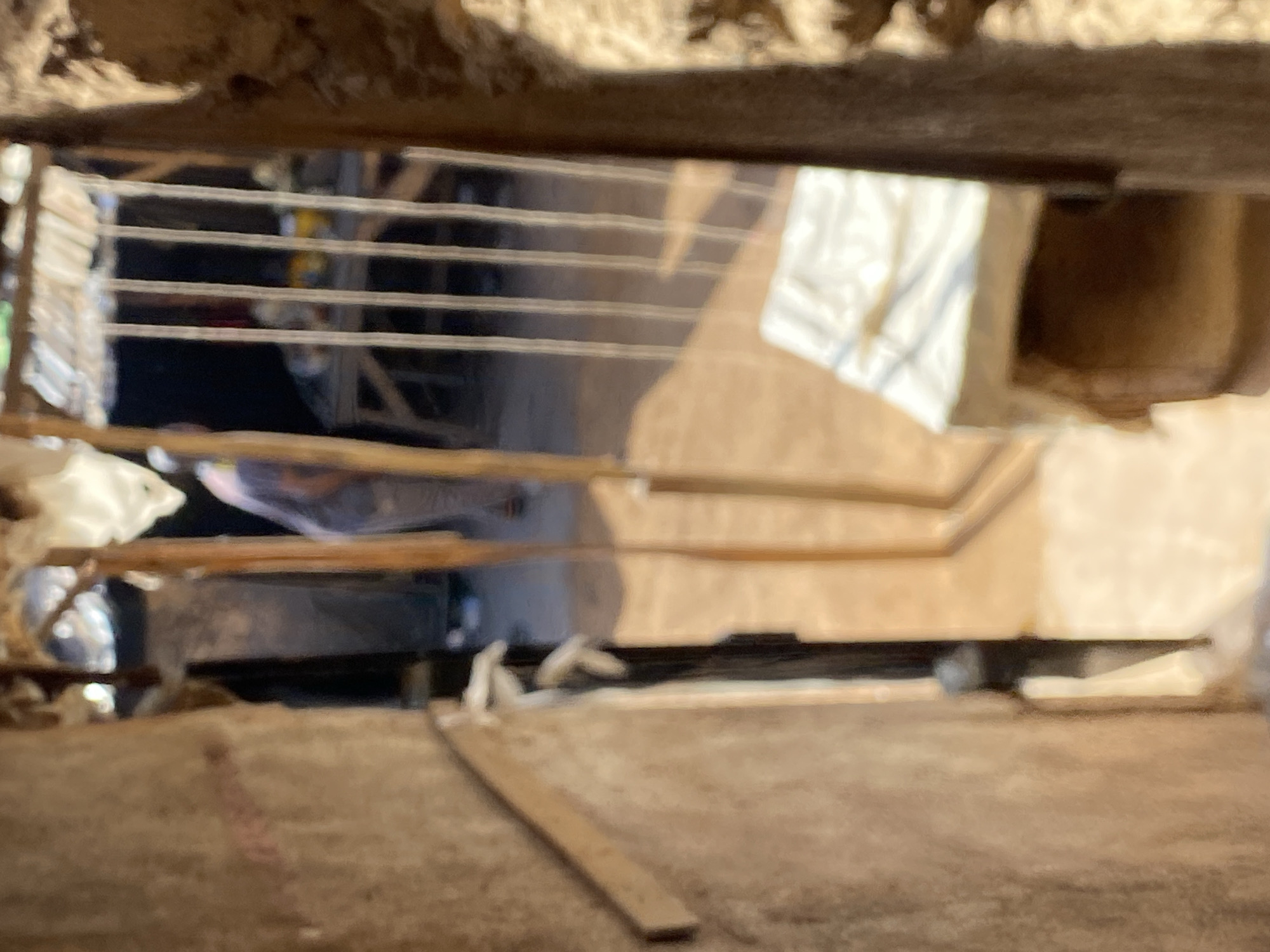 This image depicts a narrow view under a structure, possibly in Bukuru, Nigeria, given the prompt's context. A metal grill or grate spans across the opening, partially obscuring the view. Beyond the grill, various indistinct objects are visible, including what appears to be a white bag or cloth and dark, possibly mechanical, shapes. A wooden plank lies on a concrete or dirt floor in the foreground. The lighting is poor, and the image is out of focus, making precise identification of the objects and environment difficult. No people are directly visible in the frame.