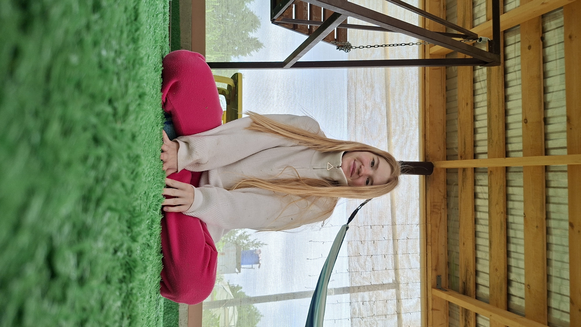 A young woman with long blonde hair is sitting cross-legged on a green artificial turf surface. She is wearing a cream-colored turtleneck sweater and bright pink pants. She is smiling and looking directly at the camera. Behind her, there is a wooden structure with a corrugated metal roof, and a hammock is visible hanging in the background. The scene appears to be outdoors, possibly in a backyard or recreational area, consistent with locations in Большой Букор, Russia.