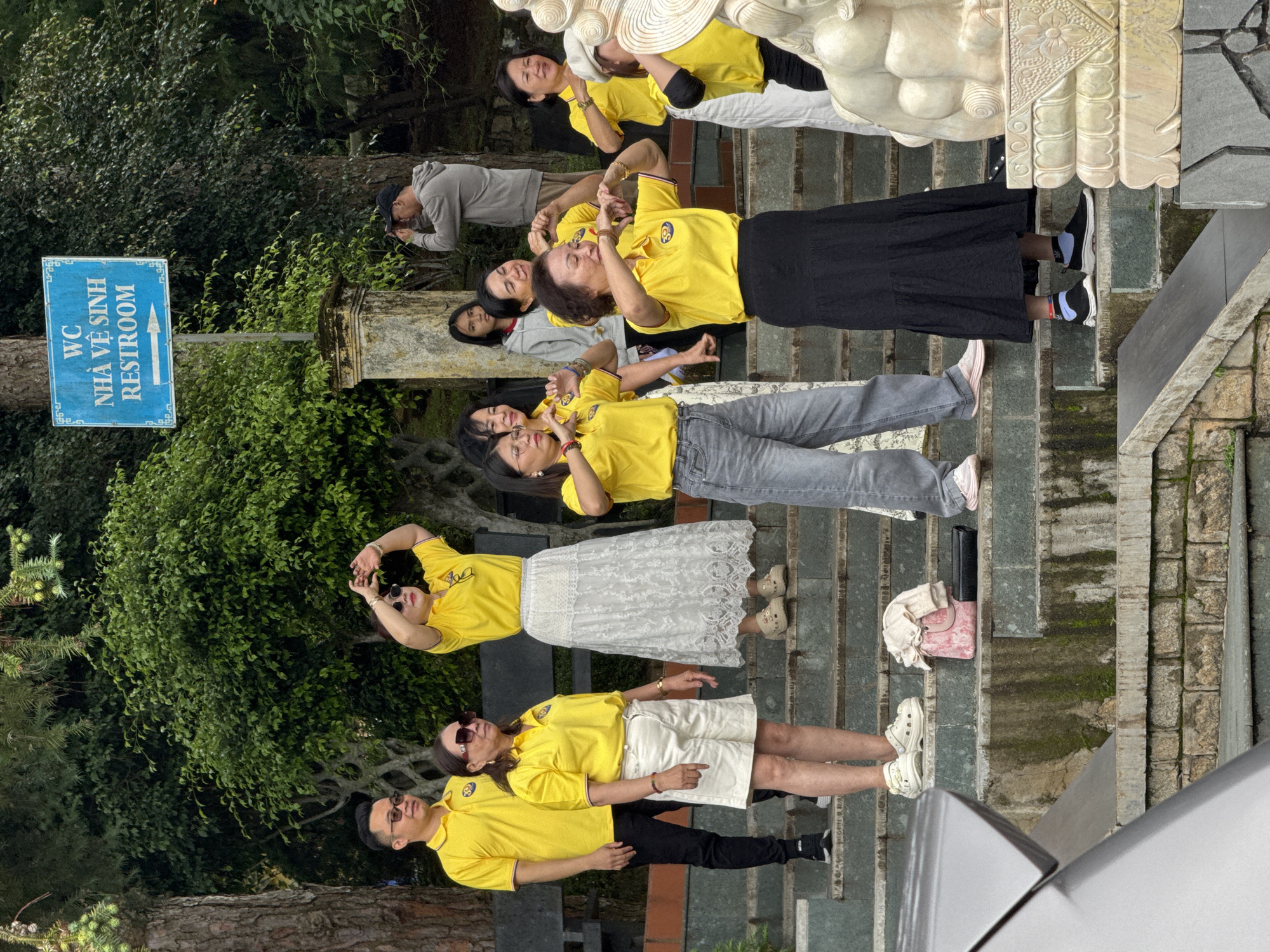 The image captures a group of people, predominantly women, dressed in matching yellow polo shirts, standing on a stone staircase outdoors. They appear to be engaged in a synchronized activity, possibly a dance or exercise, as many have their hands raised and positioned in similar gestures. The setting suggests a public space in Da Lat, Vietnam, indicated by the "WC NHÀ VỆ SINH RESTROOM" sign with Vietnamese and English text, pointing towards a restroom facility. The staircase is surrounded by lush green trees and foliage, typical of Da Lat's mountainous terrain. Several individuals are wearing sunglasses, and the overall atmosphere seems relaxed and casual. A statue, possibly of a guardian animal, is visible on the right side of the frame. The lighting suggests it is daytime.