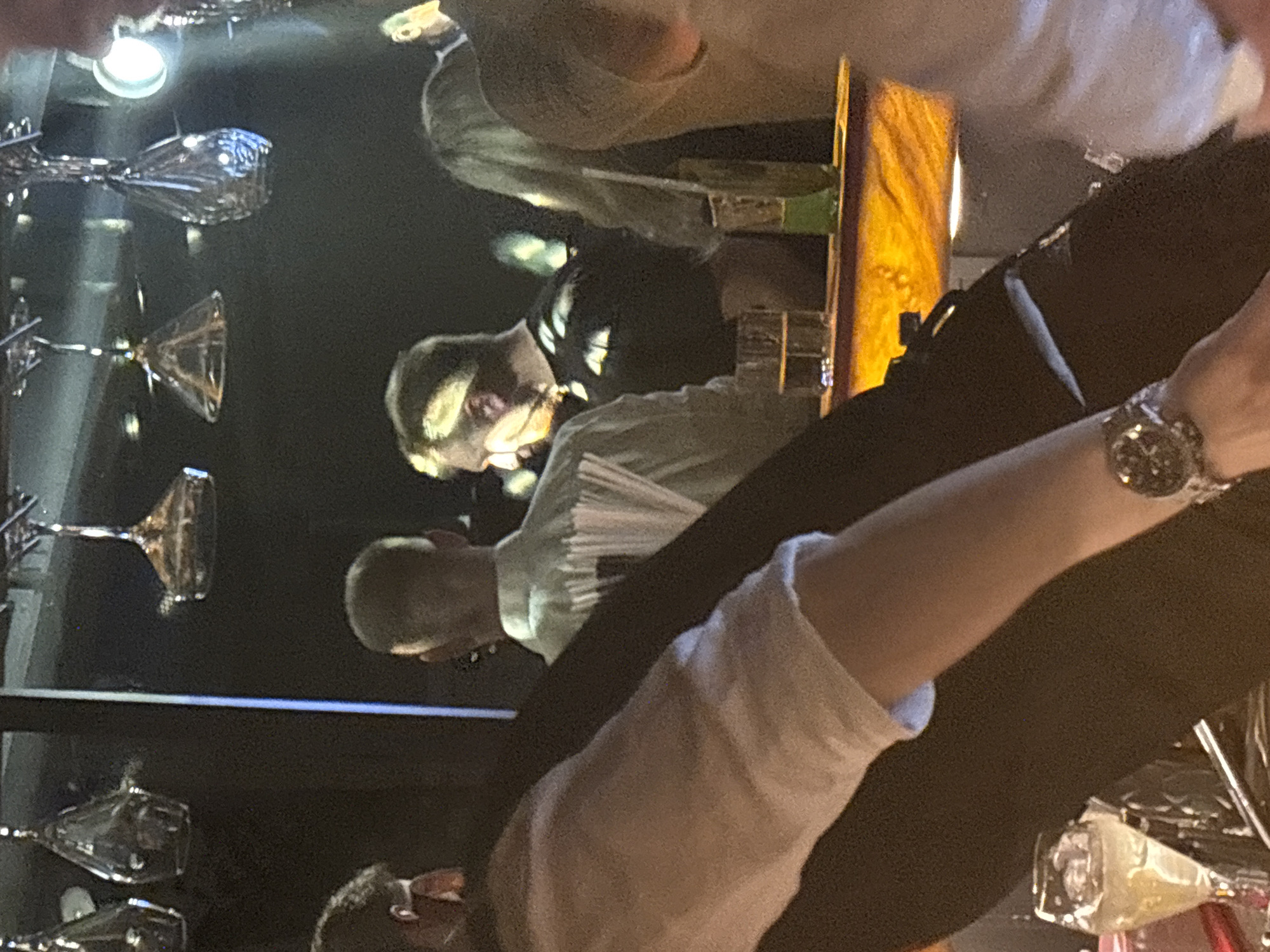 In Perm, Russia, the image captures a dimly lit scene, seemingly at a bar or restaurant. Shelves with cocktail glasses dominate the left side of the frame. Several people are present, including an individual wearing a watch. Two figures, potentially bartenders or employees, stand near a counter, with one wearing glasses. A wooden surface or counter is visible in the center. The overall impression is of a busy, upscale establishment.