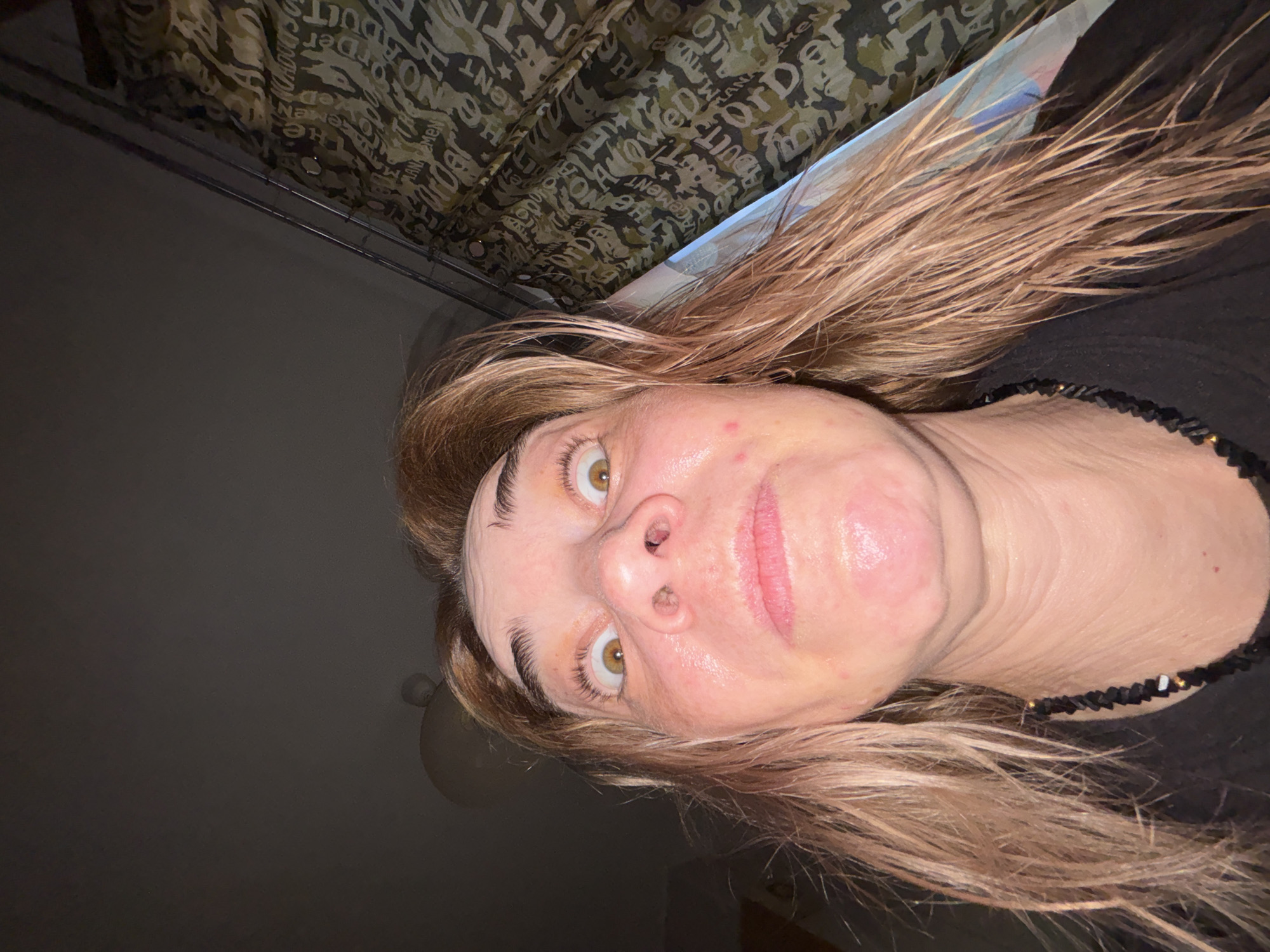 This close-up, rotated indoor portrait features an individual, likely an adult female, gazing directly at the viewer. Her light brown hair, appearing somewhat disheveled or damp, frames her face. Her skin, which shows some redness and a slightly moist or shiny texture on the forehead and nose, is illuminated by dim light. She has prominent light brown or hazel eyes and a nose piercing in her right nostril. She wears a dark top and a black beaded necklace with small lighter details around her neck.
The background is dimly lit. On one side, a dark, indistinct area is visible. Above her head, there is a patterned fabric, possibly a curtain or bedding, featuring a camouflage-like design composed of repeating, stylized text. Visible words on the fabric include "ORDER," "ADULT," "MEN," "ELEGEMENT," and "NO," presented in various orientations, including mirrored and upside down.
The low-light conditions suggest the image was taken indoors, possibly in the evening or a poorly lit room. The person's expression is neutral, conveying a candid moment. The specific location is unknown, but the context indicates Russia.