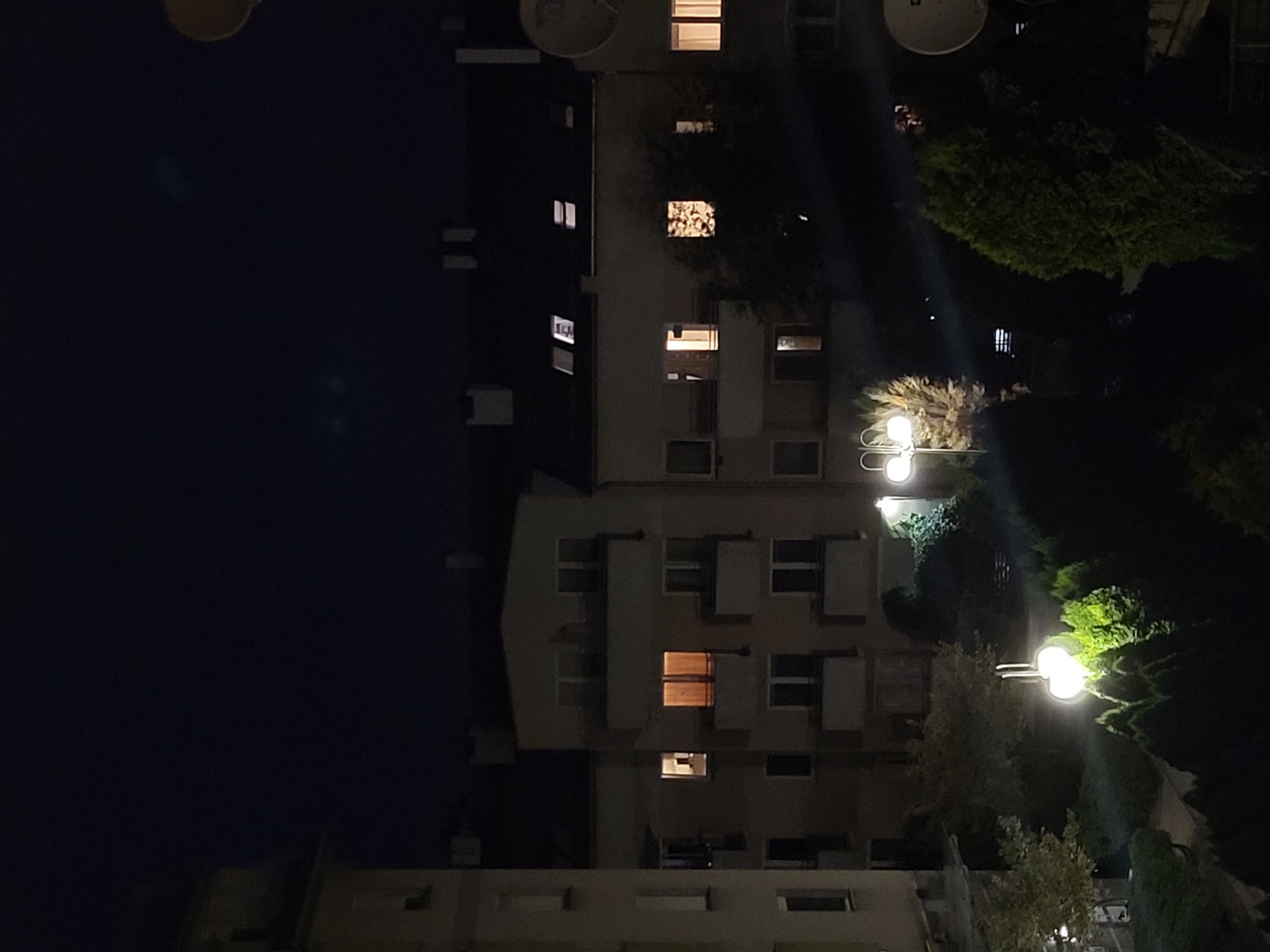 This is a nighttime, medium-distance, eye-level, wide shot of an urban residential area. The scene depicts apartment buildings with illuminated windows and trees in the foreground. The sky is dark and clear. There are no people visible in the frame. The overall impression is one of a quiet, residential neighborhood at night.