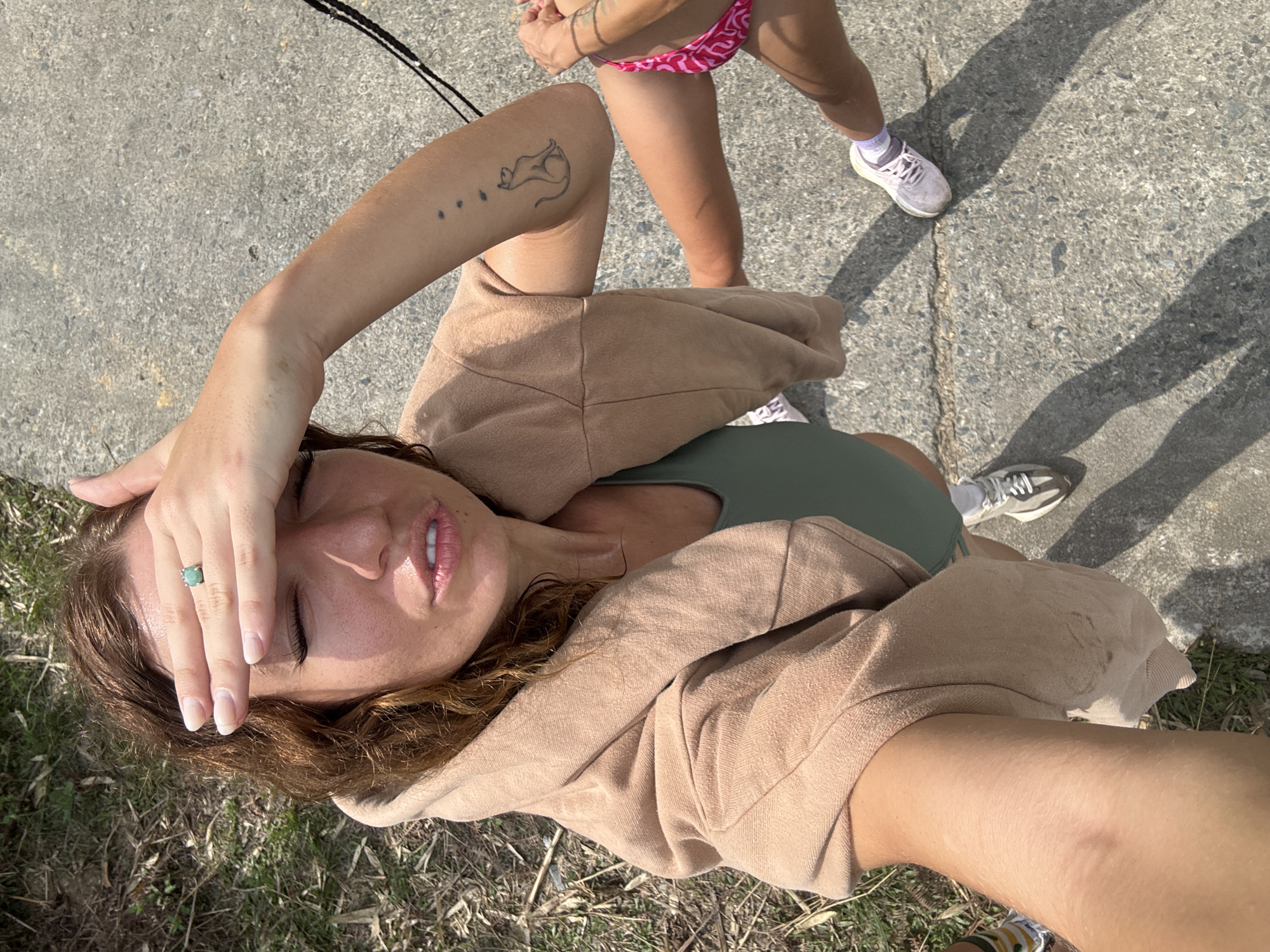 A selfie, angled downward, shows a woman's face with eyes closed and a hand on her forehead wearing a turquoise ring. She has brown hair, fair skin, and is wearing a green top and a tan hooded jacket. The background includes a concrete surface, a second person wearing a pink patterned bathing suit bottom, and partial views of shoes. There is also grass and shadows visible. The location is likely outdoors, with a mix of pavement and grassy areas. It is unlikely to be Suối Giàng, Vietnam, given that bathing suits are not commonly worn in that area of Vietnam.