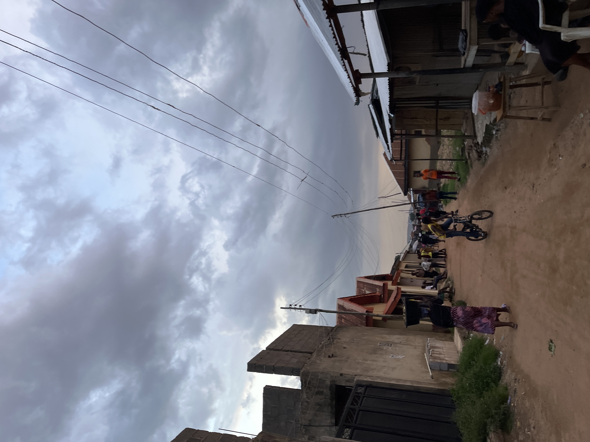 The image depicts a street scene in Jos, Nigeria, under an overcast sky, suggesting it is either early morning or late afternoon. The foreground features a dirt road with several people scattered throughout. A person is riding a bicycle, and another individual in a striped skirt carries a large object on their head. In the background, buildings with corrugated iron roofs and unfinished construction are visible. Some structures appear to be shops or homes. A few individuals are gathered near a table and chair setup outside one of the buildings. The overall atmosphere seems to be a typical day in a residential or commercial area, with people engaged in various activities. The lighting is dim, consistent with a cloudy day. No visible text is present in the image.