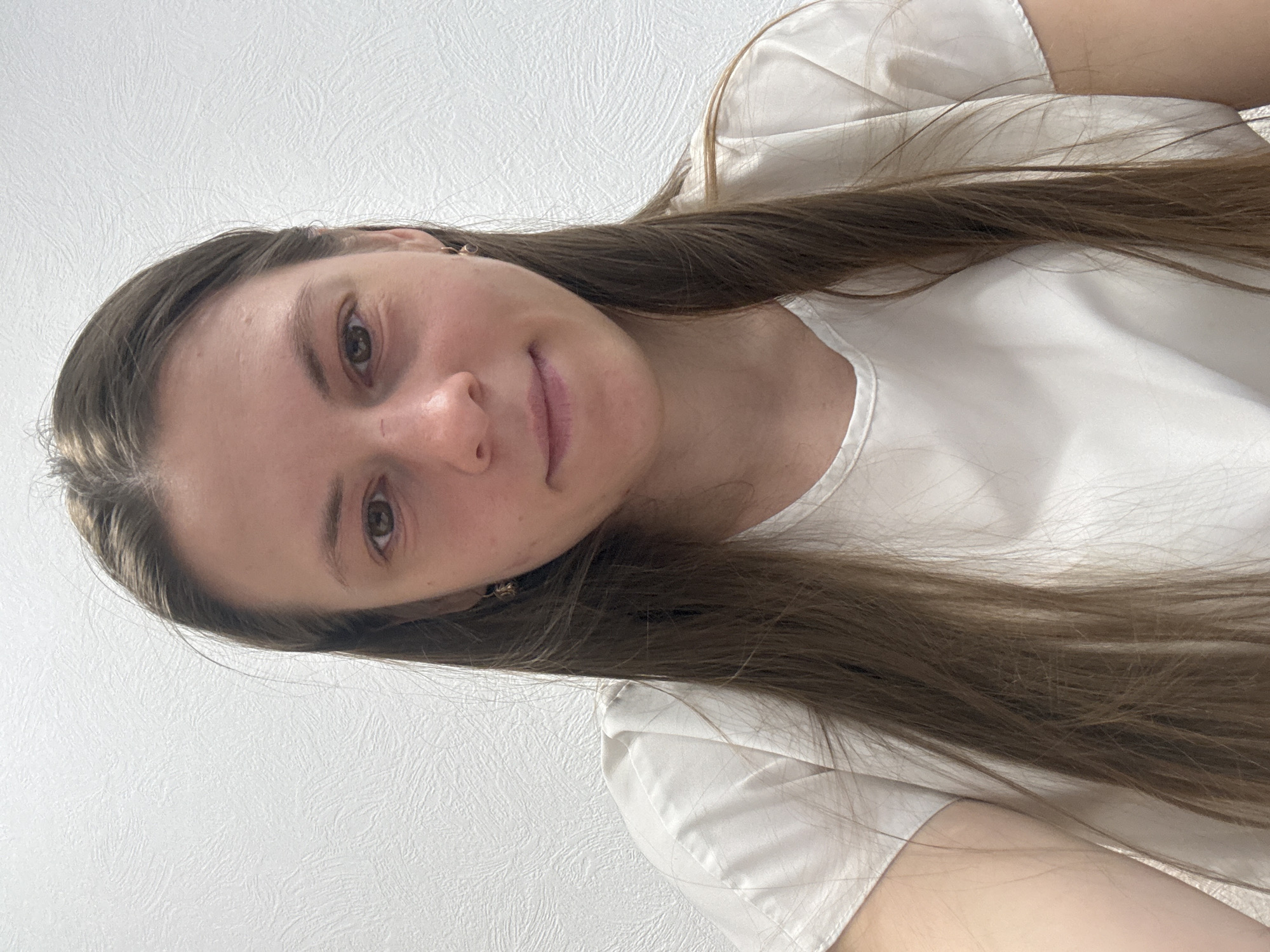 A woman with long, brown hair and fair skin is facing the camera. She is wearing a white, collared shirt. The background is a white wall. There is no information to determine the location in the city of Chaikovsky, Russia.