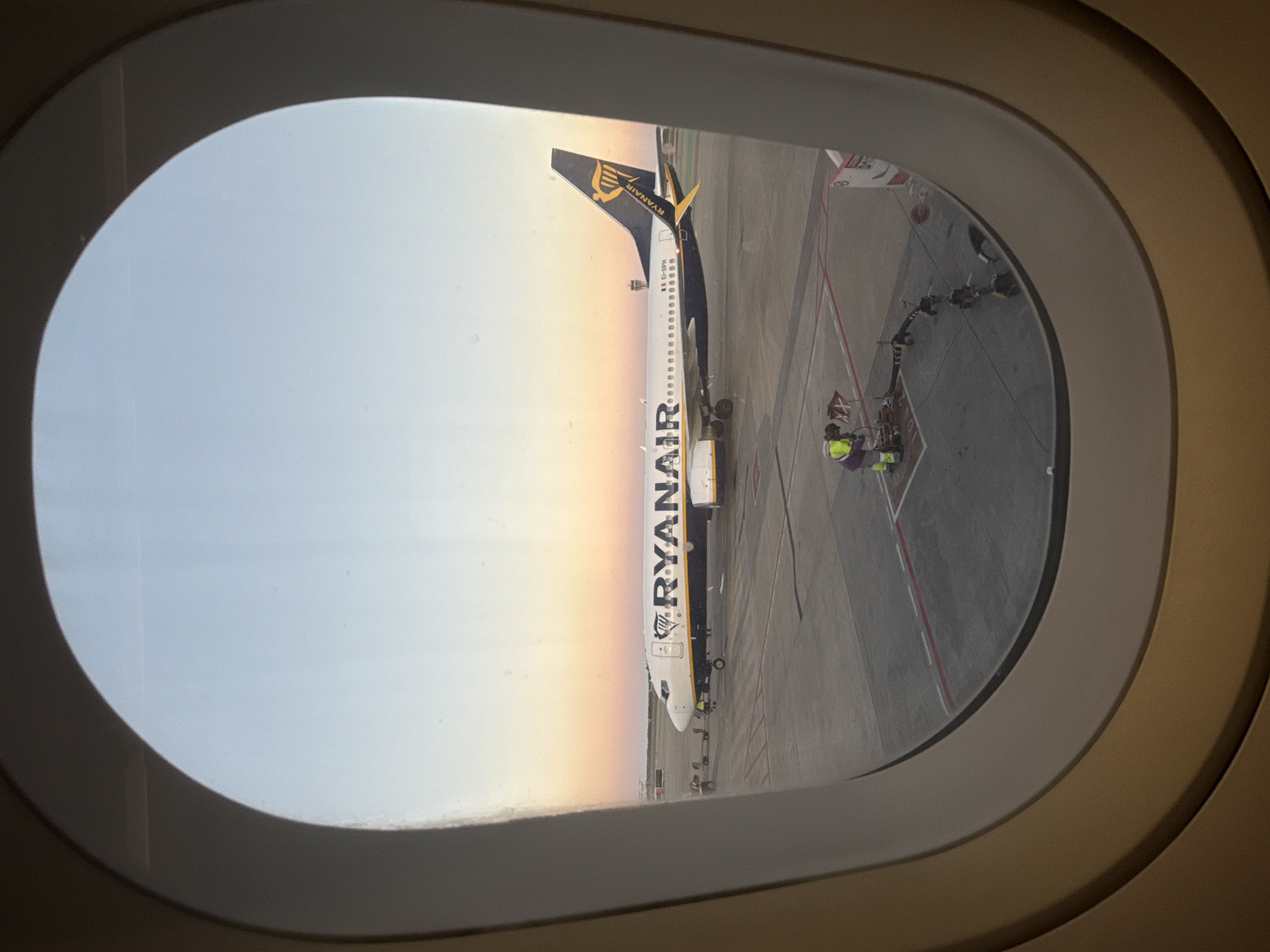 An airplane, identified by its "RYANAIR" livery, is viewed through an airplane window. The plane is parked on a tarmac near what appears to be a jet bridge. Personnel are near the aircraft. The sunset suggests the setting is either dawn or dusk. This is likely at or near the city of el Prat de Llobregat, Spain, due to Ryanair’s operations there.