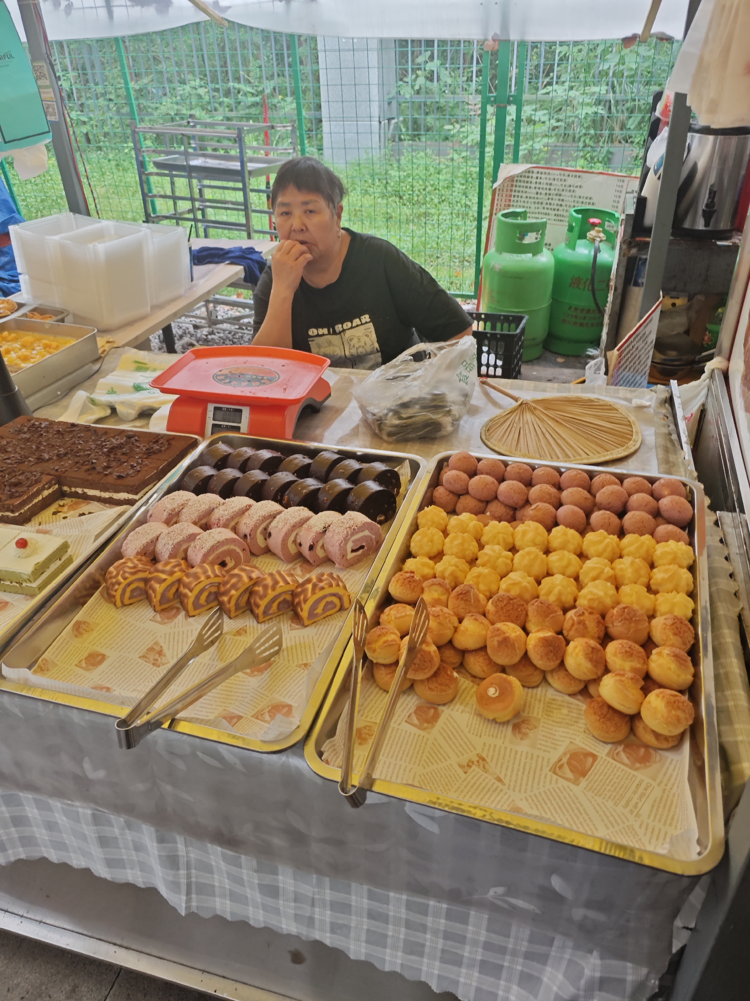 A middle-aged Asian woman is sitting at a table covered with trays of baked goods at an outdoor market. She is wearing a dark t-shirt and has short, dark hair. She is looking at the camera with a neutral expression and has her fingers to her mouth.
On the table in front of her, there are four large metal trays filled with pastries, cakes, and other baked goods. The pastries are of various colors and shapes. There is also a red scale, clear plastic food containers, a plastic bag, and a woven fan on the table. Behind her, there are green gas tanks. A metal frame structure is visible in the background through a green fence. The location is Shapingba District, China.