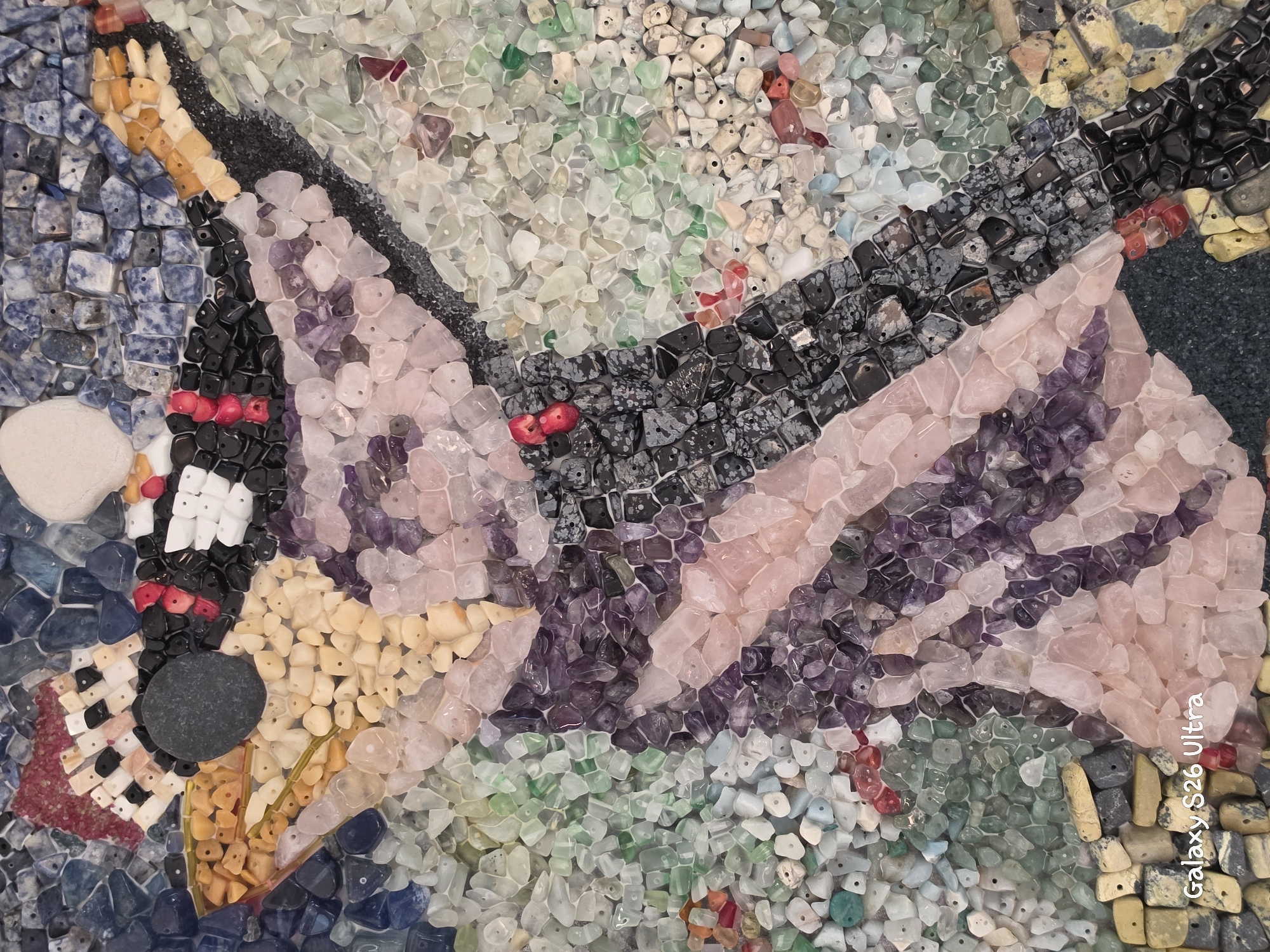 The image is a close-up, overhead shot of a mosaic artwork composed of small, polished stones and beads. The mosaic depicts a stylized figure, possibly an animal like a giraffe or a bird, with a dark circular stone representing an eye. The figure has a patterned body and head, created with contrasting colors and textures of stones such as purple amethyst, pink rose quartz, light green aventurine, black obsidian, and yellow and beige stones. The background is a mix of blue, grey, and lighter colored stones. In the bottom left corner, the text "Galaxy S26 Ultra" is visible, indicating it is a photograph taken with that device.
There is no information in the image that indicates it is located in Plovdiv, Bulgaria.