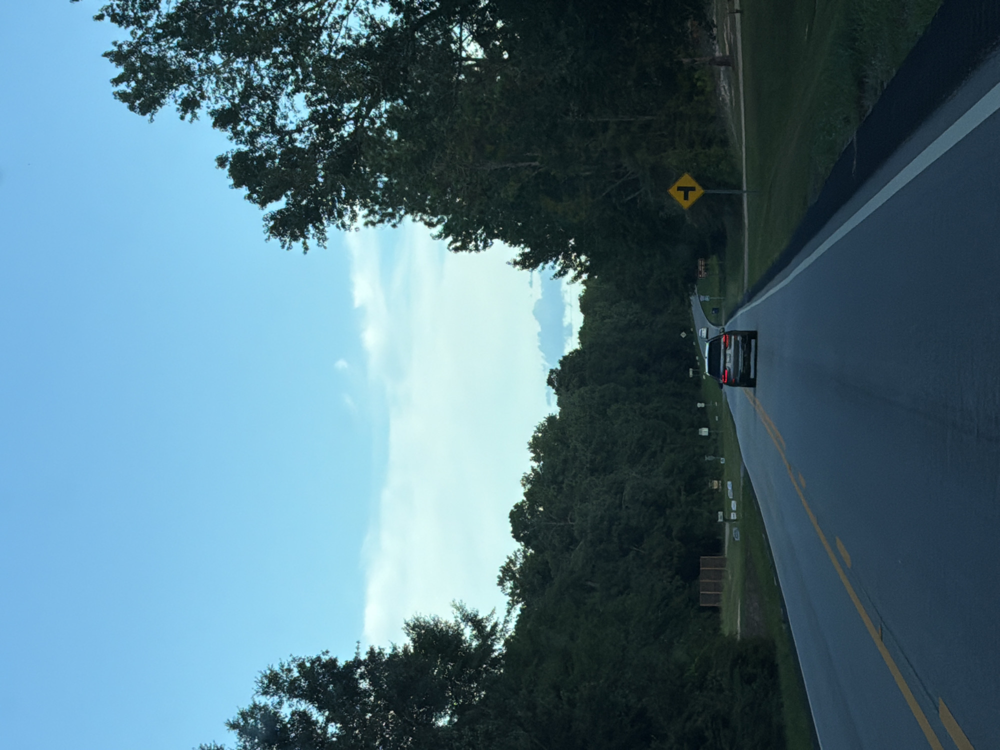 The image shows a two-lane road with a yellow dividing line and a white outer line, likely in Andalusia, United States, given the information provided. The road is flanked by dense green trees on both sides, creating a tunnel-like effect. The sky is a clear blue with some wispy white clouds. A black SUV is driving away from the camera in the right lane, while another car is visible further ahead on the left lane. A yellow traffic sign indicating a T-junction is positioned on the right side of the road, near the edge of the tree line. The overall impression is of a scenic drive through a wooded area, possibly in a rural or suburban setting.
