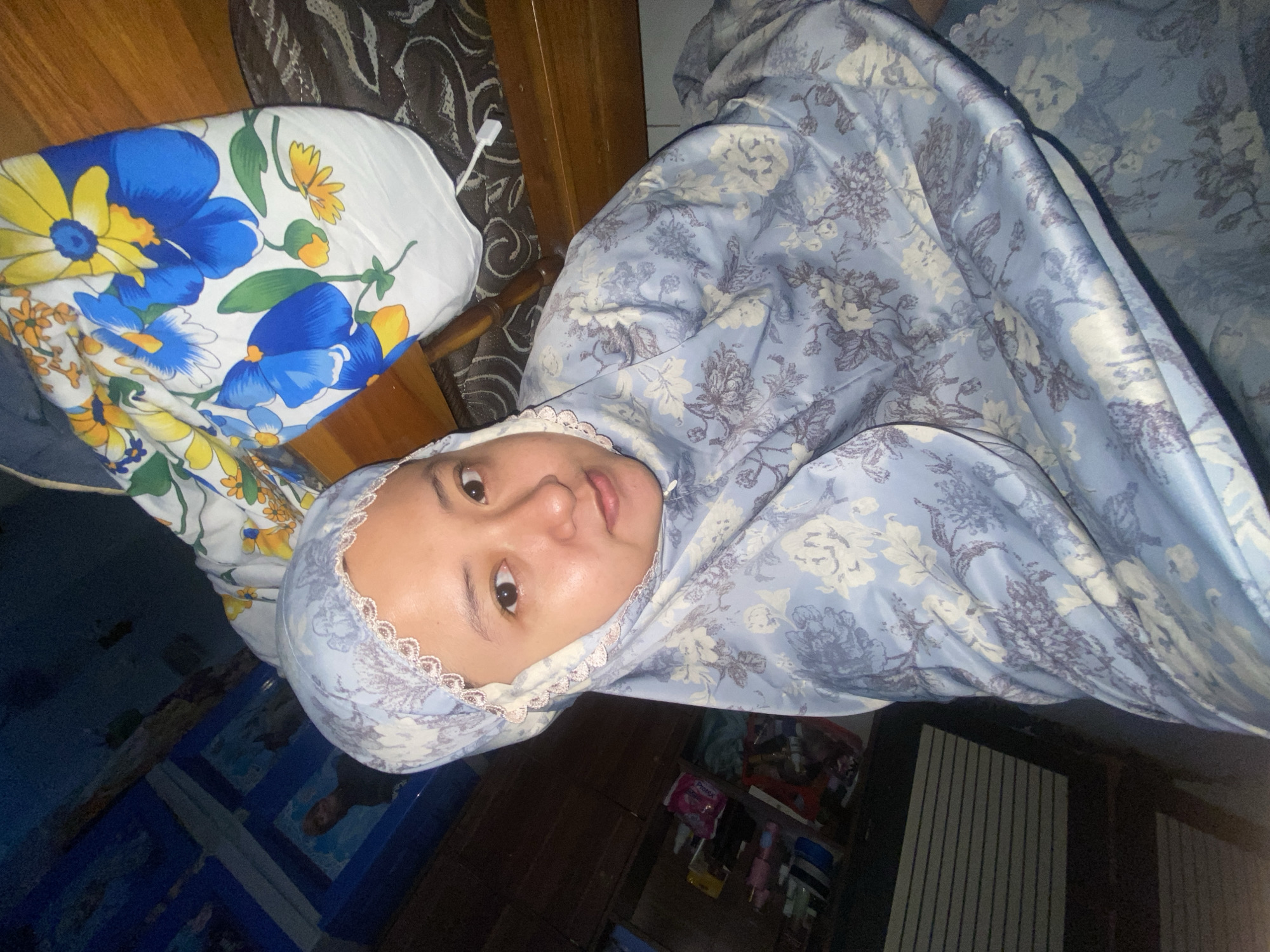 A woman in a light blue, floral prayer garment is pictured from a slightly elevated angle in what appears to be a domestic setting, possibly a bedroom. The background shows a wooden furniture item and a patterned fabric, possibly a blanket or a large cushion with blue and yellow floral designs. To the left, a dark wooden cabinet with shelves is visible, containing various items, including what seem to be toiletries and other personal belongings. The overall lighting is dim, suggesting an indoor environment. The specific location in Gelam, Indonesia, cannot be determined from the visual information provided in the image.