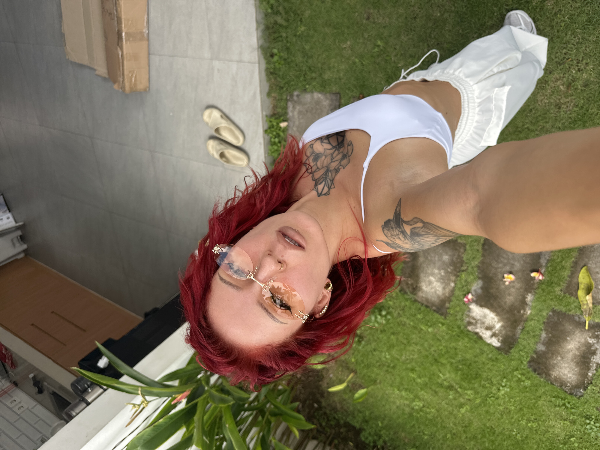A selfie of a woman with red curly hair, tattoos on her chest and arm, wearing sunglasses, a white top and pants, and standing on a patio next to a grassy area. There are sandals and a box on the patio. The location is in North Kuta, Indonesia.