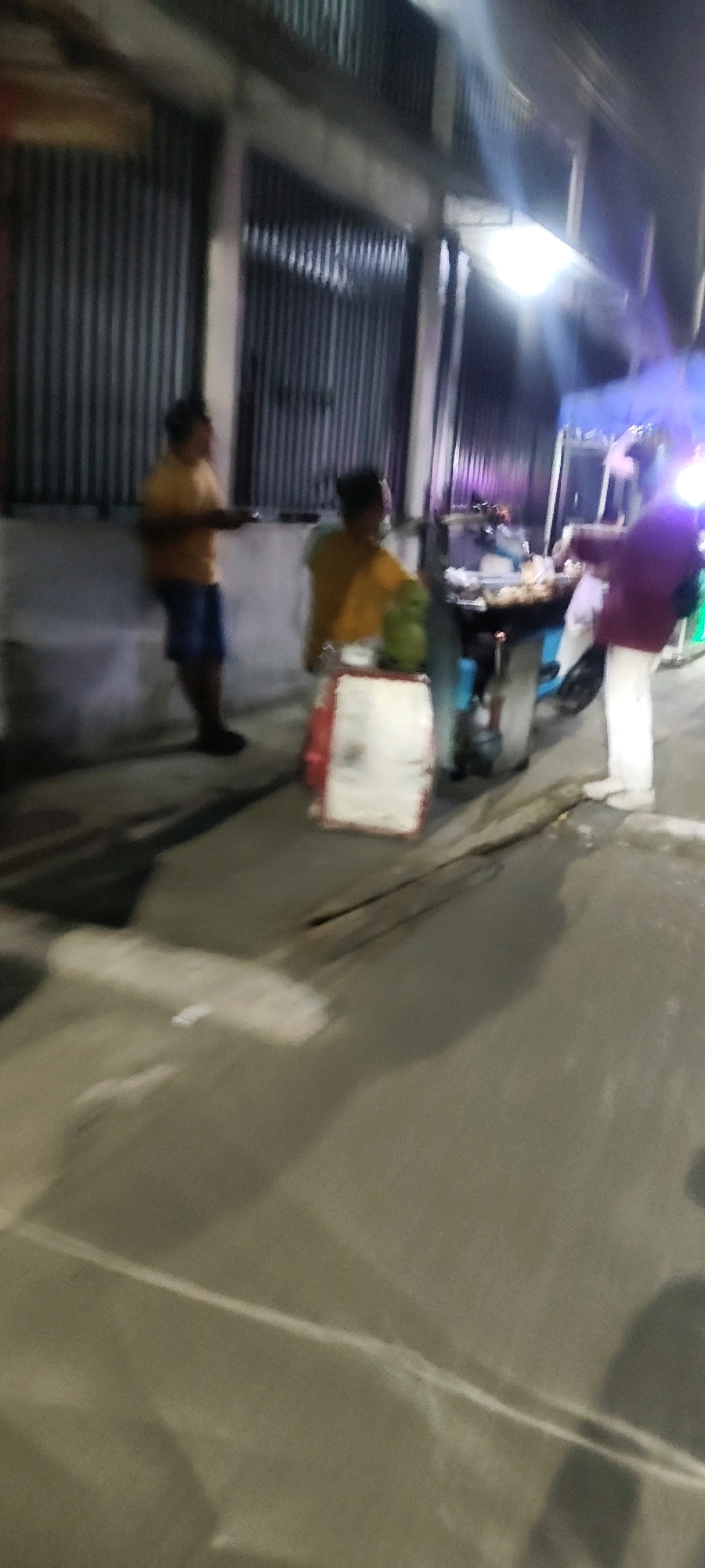The image depicts a dimly lit outdoor scene, likely at night, in Mekarmukti, Indonesia. In the foreground, a paved surface with some debris and a visible crack is present. A shadow of a person is cast on this surface.
The midground features a street food stall. A person wearing a burgundy top and white pants stands near the stall, facing away from the camera. Another individual, wearing a yellow shirt and blue shorts, stands to the left of the stall, holding a plate or tray. A third person in a yellow shirt is partially visible behind the stall, near a green cylindrical object. The stall itself has a cart with a display of food items. A red and white cooler or container is positioned in front of the stall.
The background consists of a corrugated metal wall, likely part of a building or structure, with some overhead lighting illuminating the scene. A blue tent structure is visible further back, suggesting a market or event setting. The overall atmosphere is that of a typical street food vendor operation at night.