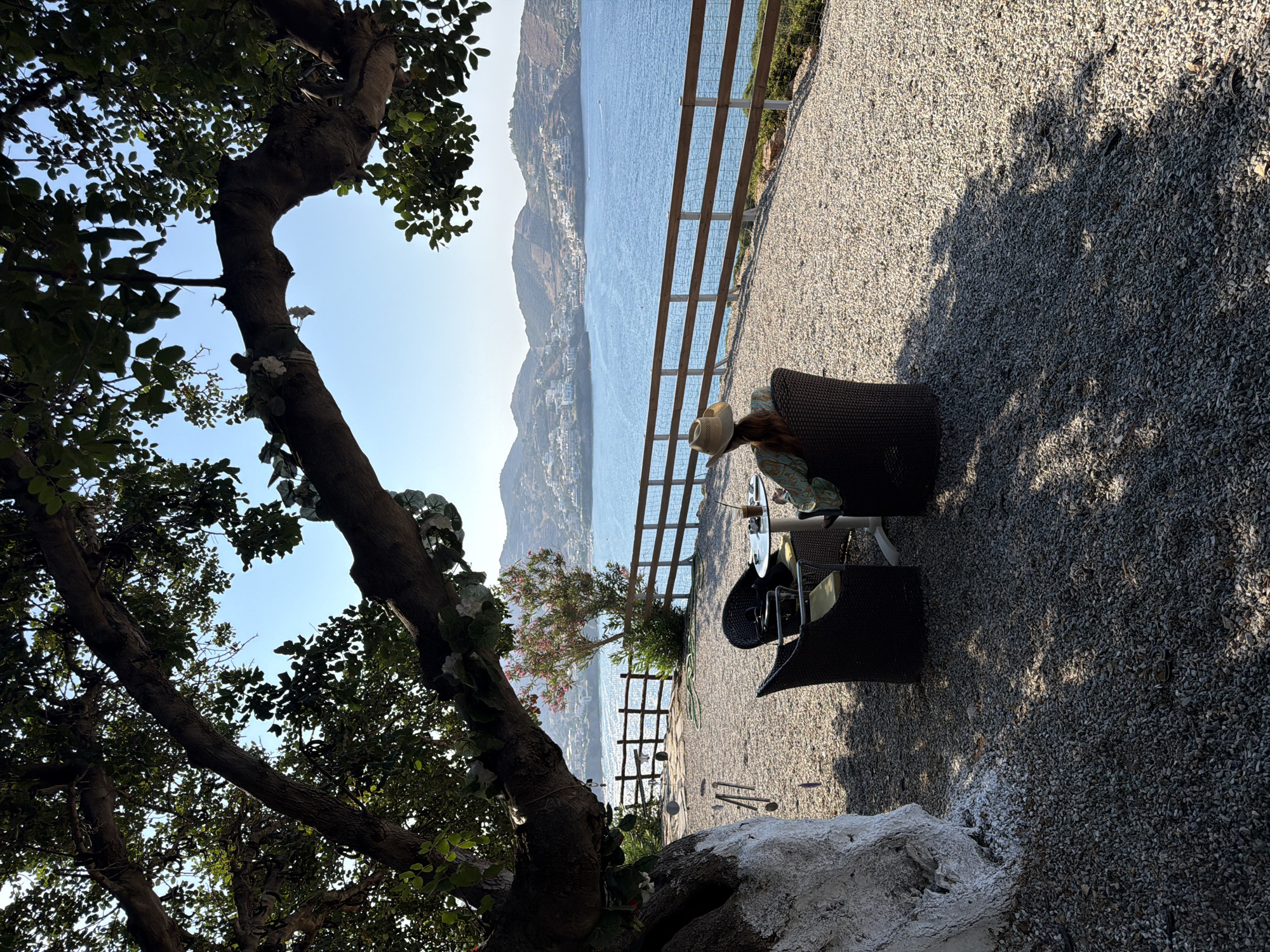 In a seaside cafe in Bali, Greece, a woman with a straw hat sits at a table by the water, enjoying a drink. The cafe is situated on a gravel patio under a large tree with drooping branches. A wooden fence separates the patio from the shimmering blue sea, beyond which lies a town nestled on rolling hills. The sky is clear and blue, and the sun casts dappled shadows on the ground. Two wicker chairs and a glass-topped table with drinks are visible on the patio, suggesting a tranquil moment of relaxation with a scenic view.
