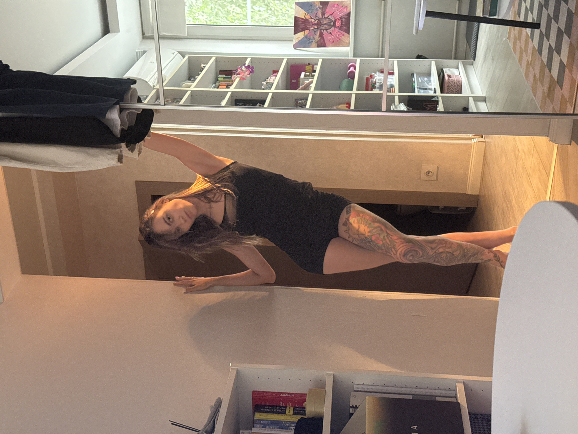 A woman is performing a yoga pose, possibly a side plank or a variation of it, in a room. She is wearing a black dress. Her body is extended horizontally, supported by one arm, while her legs are also extended. She has tattoos on her legs and arms, including intricate designs on her left leg. Her hair is long and brown, falling around her face.
The background reveals a domestic setting. There are shelves filled with various items, including what appear to be books and decorative objects. A window with greenery visible outside suggests daylight. A patterned rug is visible on the floor. The overall lighting indicates it is daytime. The woman's expression is focused, suggesting concentration on her pose. The scene appears to be indoors, likely a home or studio in Moscow, Russia. No text is clearly legible in the image.