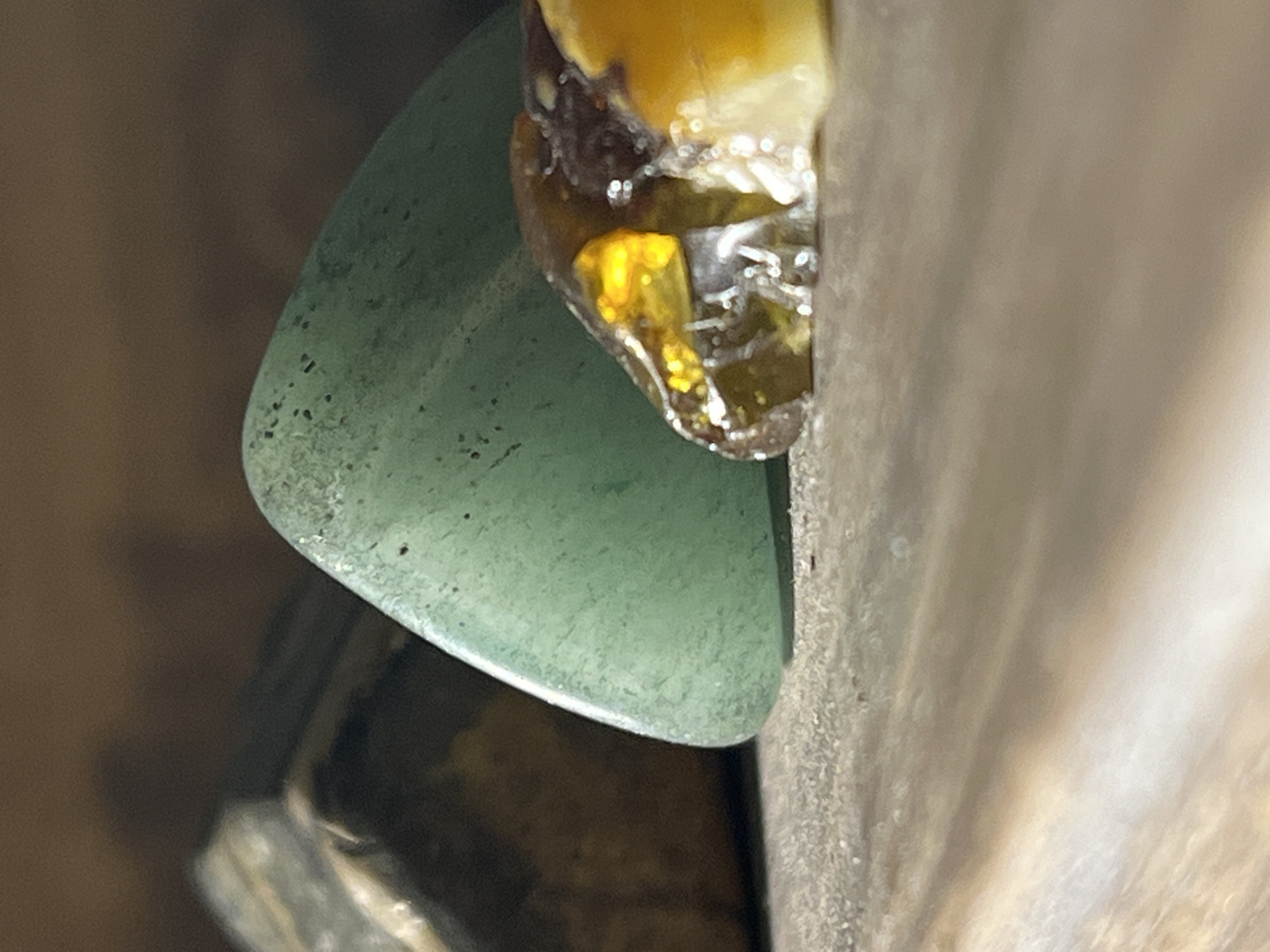 A close-up shot captures a green, possibly aventurine, gemstone resting next to a brown and amber colored object, likely amber or a resin, touching a light brown surface. The setting suggests an indoor environment with additional partially visible objects in the background. The image offers no contextual clues to identify the location as Unknown, Unknown.