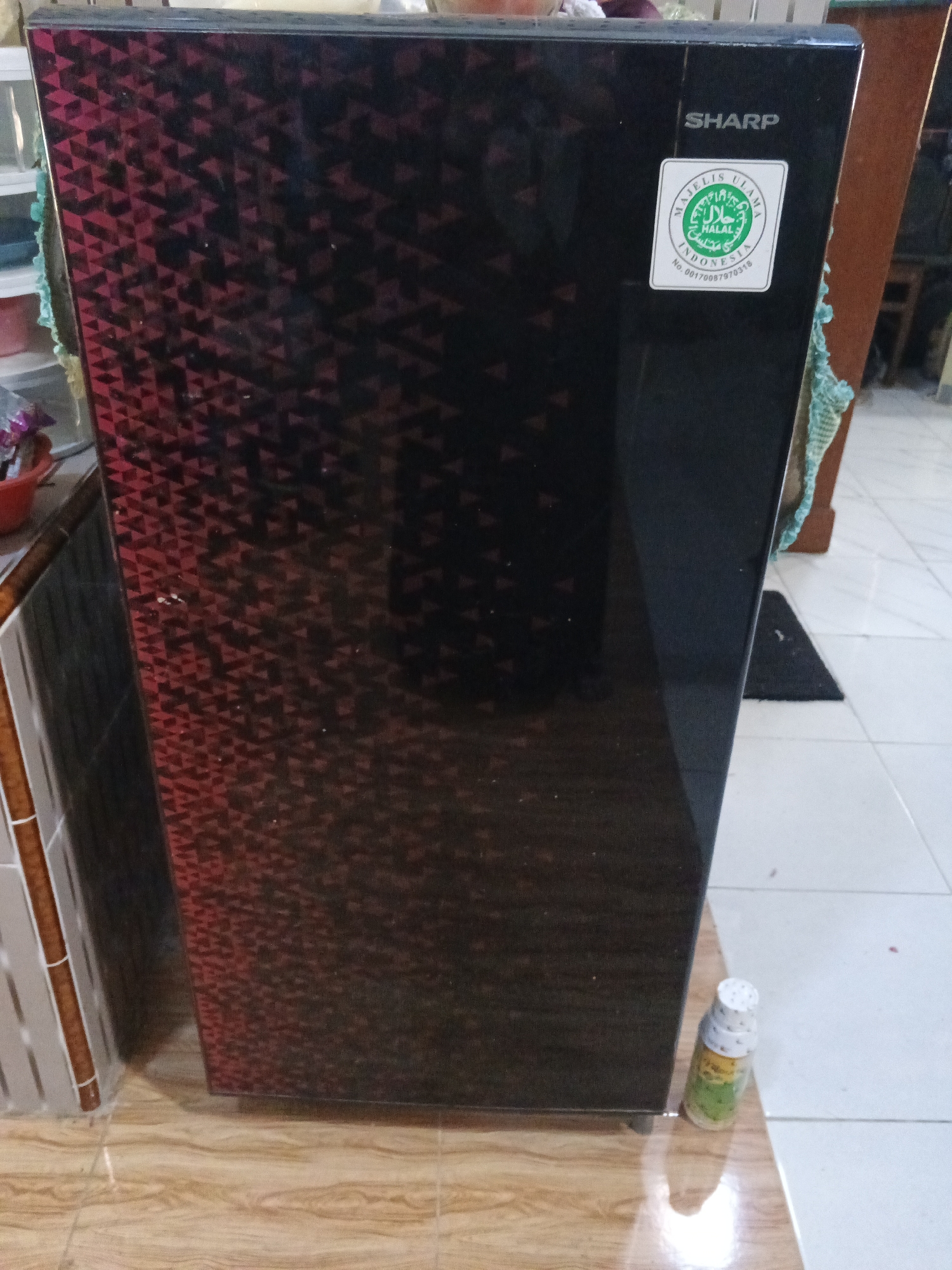 A black Sharp refrigerator with a red triangular pattern on its door is standing on a tiled floor in Kemangsen, Indonesia. A green and white Halal sticker is affixed to the upper right portion of the refrigerator door. A small bottle of liquid is on the floor to the right of the refrigerator. The background includes a wooden chair and cabinets.