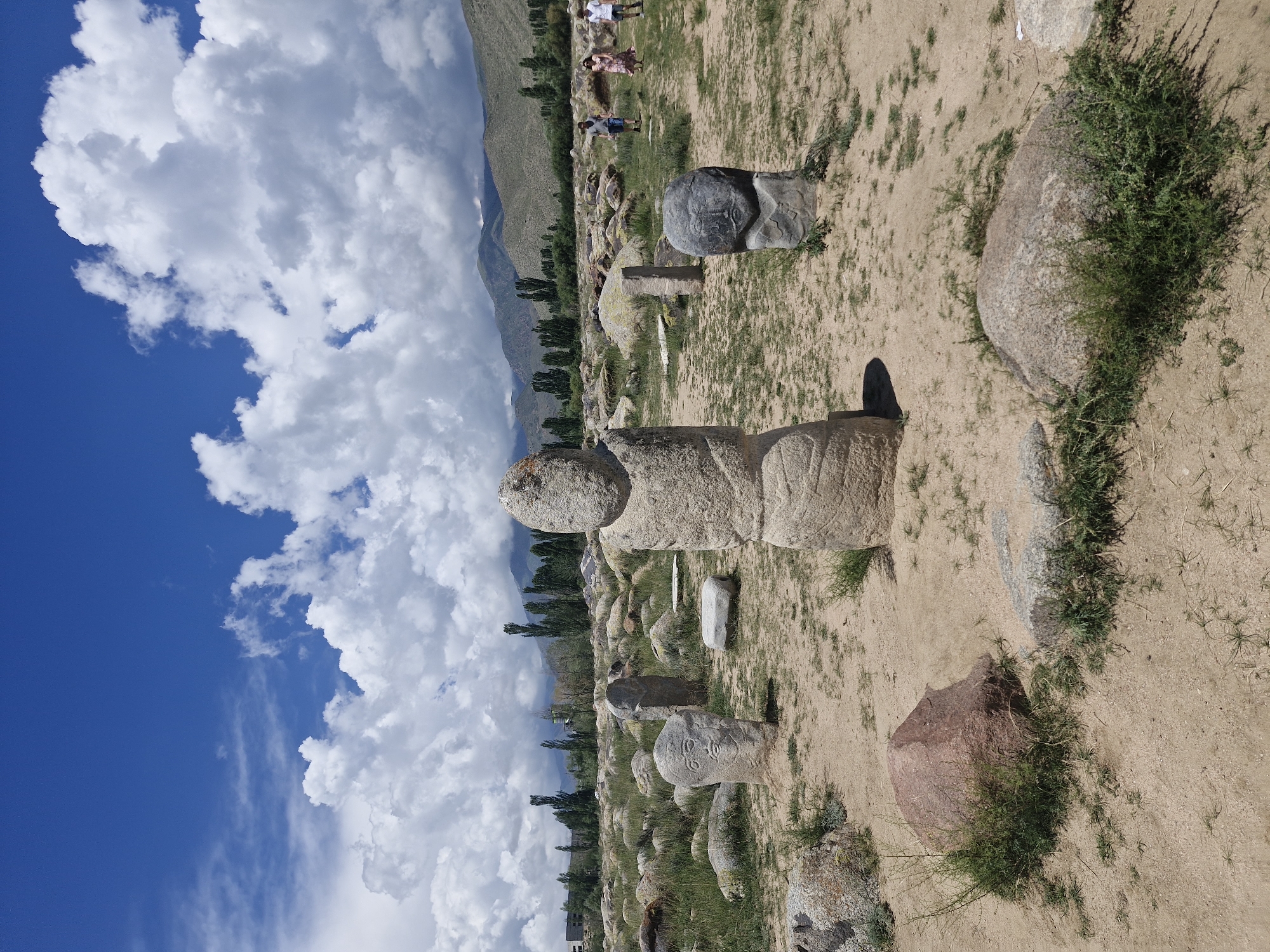 The image depicts a historical outdoor museum with ancient stone sculptures, likely petroglyphs, located in Cholpon-Ata, Kyrgyzstan. The sculptures, varying in size and form, are scattered across a sandy, open field dotted with rocks and sparse vegetation. Some of the stones are carved with faces and patterns. In the background, a line of trees and distant mountains under a bright blue sky with white cumulus clouds complete the scenic environment. Three individuals, two adults and a child, are walking in the middle ground of the scene. The overall impression is one of an archaeological site preserved in a natural landscape.
