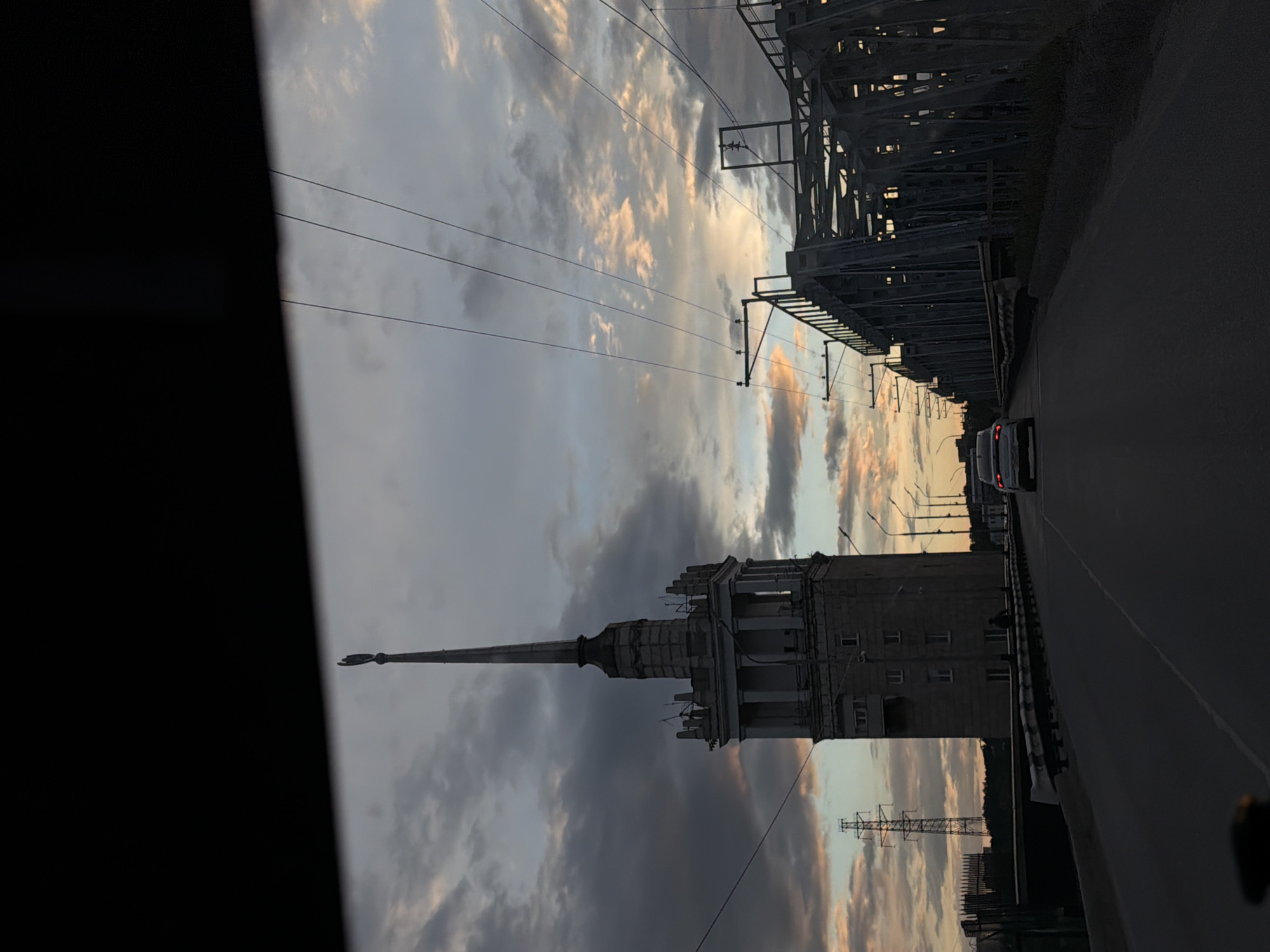 The image depicts a view from a vehicle traveling on a bridge in Perm, Russia. The most prominent landmark is the Perm Railway Administration building with its distinctive spire. Overhead are power lines and a railway bridge structure. A car is visible on the road ahead. The sky is filled with clouds, tinged with the colors of sunset. The left side of the frame is obscured by part of the vehicle's interior.