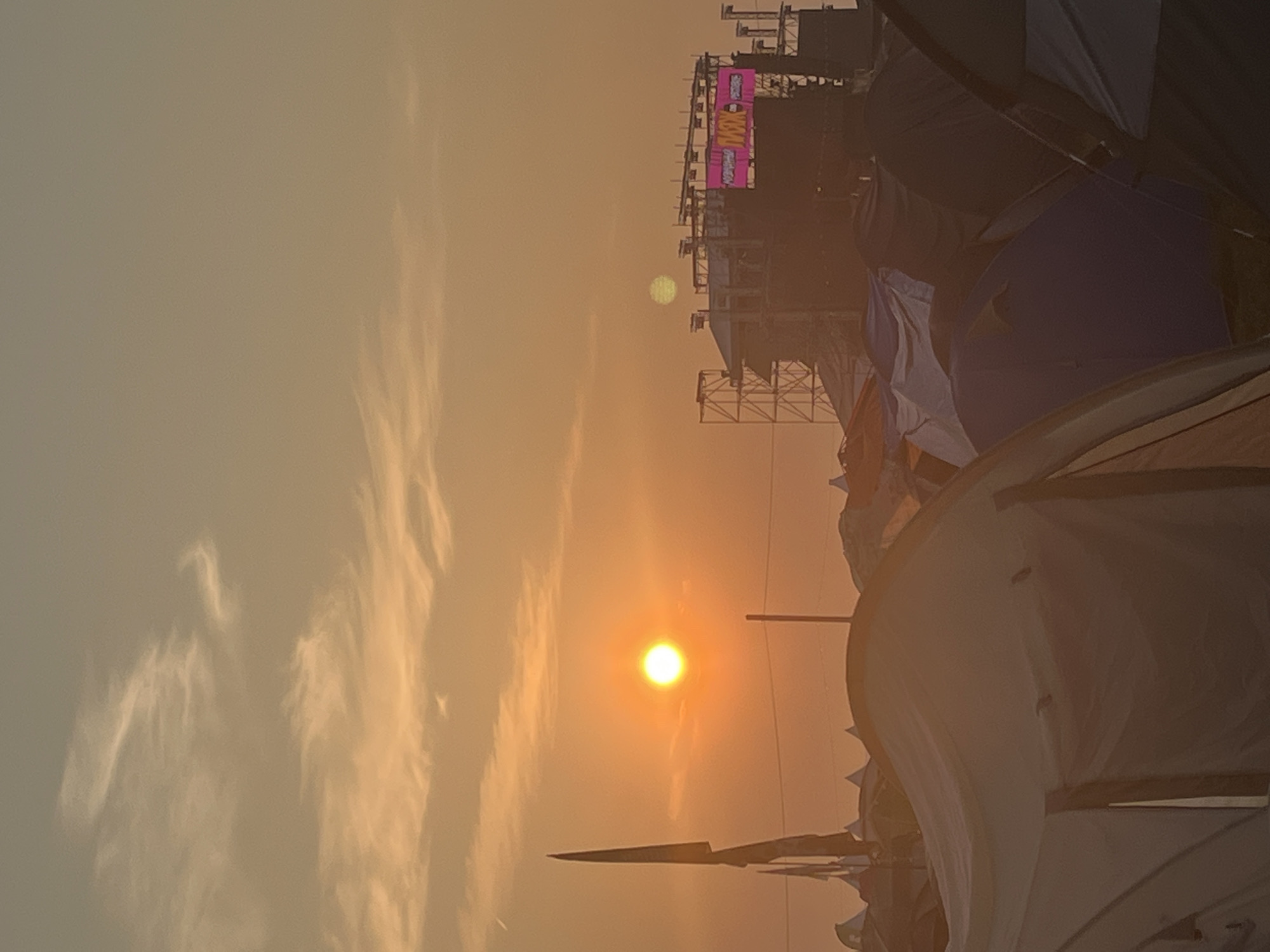 This is a vertical, eye-level, medium shot of a festival campsite at sunset, with a stage in the background, taken in Surakovo, Russia. The sun, a bright orange orb, is low in the sky, casting a warm glow over the scene. Wispy clouds are scattered across the sky. In the foreground, numerous tents are pitched, their fabric illuminated by the setting sun. Some tents are light-colored, while others are darker shades of blue and purple. A tall, black flag with a pointed tip stands to the left of the sun, partially obscuring it. Behind the tents, a large stage structure is visible, with scaffolding and large screens. A pink banner with text in Cyrillic is displayed on the screen, one of which reads "MAX" in large letters. The overall impression is one of a vibrant outdoor event nearing its end for the day.