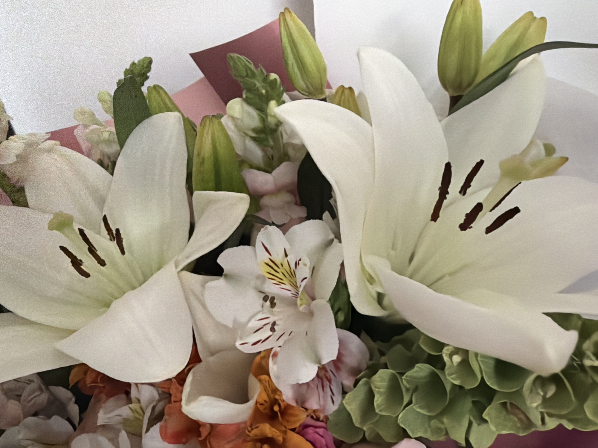 A close-up photograph shows a bouquet of flowers. Predominantly white lilies are featured prominently with brown stamens. There is also an Alstroemeria flower visible which is white with yellow and brown markings. Other floral elements include green buds, foliage, and blossoms in shades of pink, orange, and white. There is also pink and burgundy packaging material evident. The background is plain white.