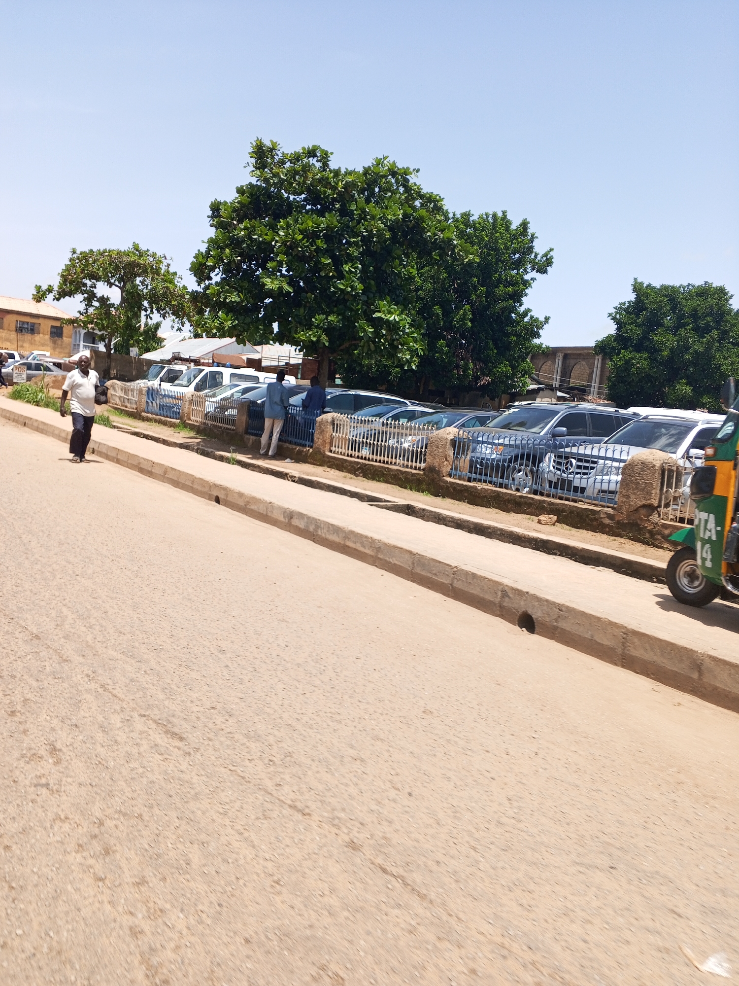 In New Stadium, Nigeria, on a bright, clear day, a street scene unfolds. A beige asphalt road occupies the foreground, leading towards a raised sidewalk made of concrete. Beyond the sidewalk, a row of parked cars, primarily white vans and dark sedans, are visible behind a metal fence. Several trees, lush with green foliage, provide shade along the sidewalk. A man in a white polo shirt and dark pants walks away from the camera on the sidewalk. To the right, a green and yellow auto-rickshaw, with the partial text "TA-14" on its side, is parked or moving slowly. Buildings, including one with an orange facade, are visible in the background. The overall impression is a typical urban street in Nigeria on a sunny day.