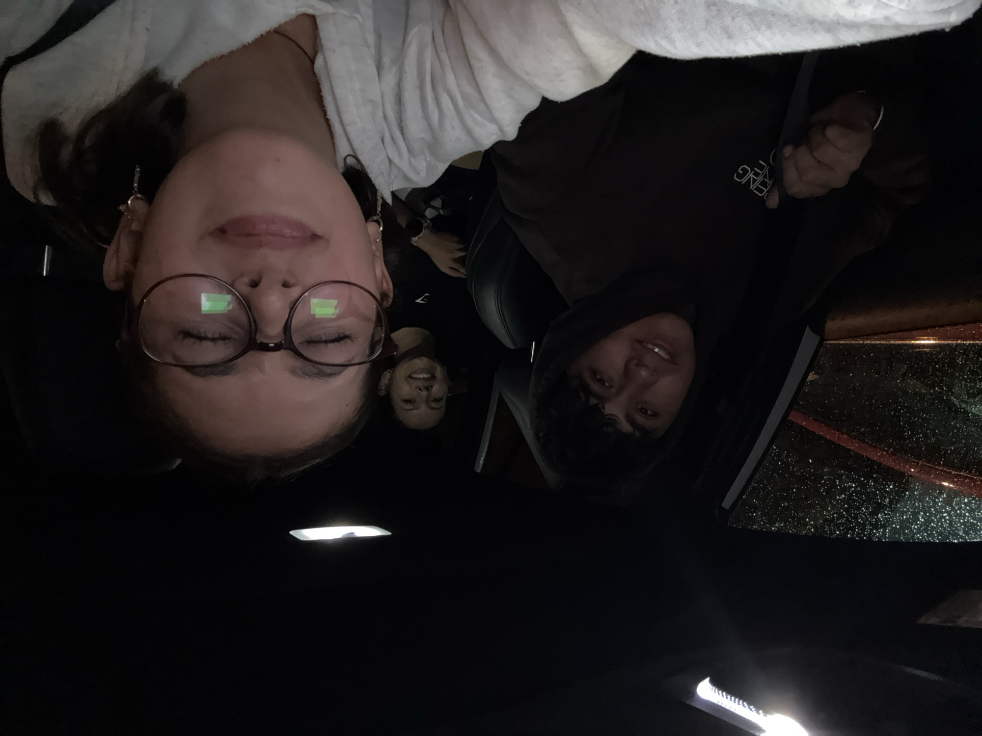 Three individuals are depicted in the interior of a vehicle. The person in the foreground on the left has glasses and is wearing a light-colored top. To the right of them, another person is wearing a dark-colored hoodie. A third person is visible in the background, between the two in the front. Rain is visible on the car window. The image was taken inside of a car, at night, in San Andrés Cholula, Mexico.