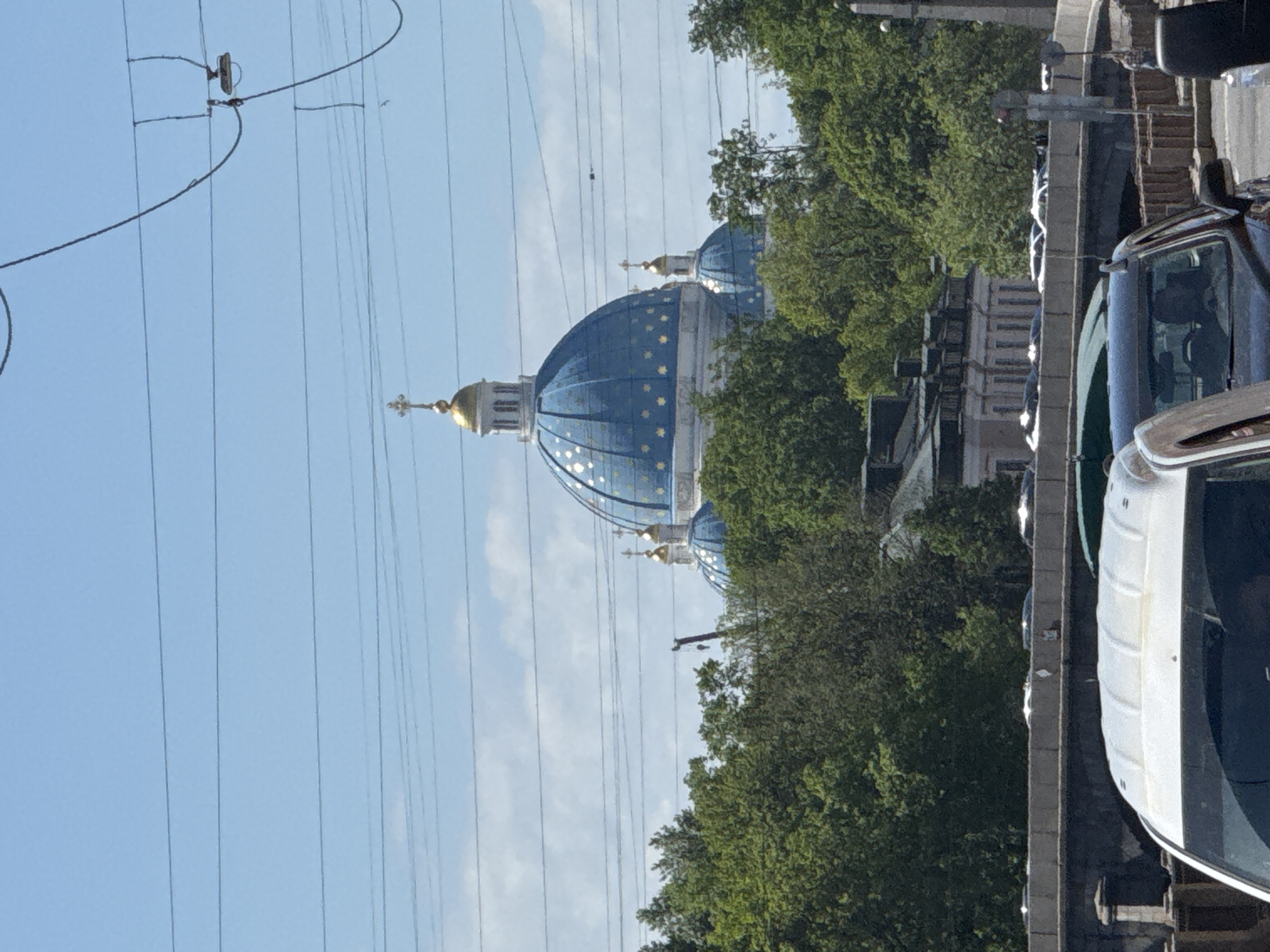 The image shows a partial view of the Trinity Cathedral in Saint Petersburg, Russia, recognizable by its distinctive blue domes adorned with golden stars. The cathedral is partially obscured by green trees. Overhead are numerous power lines stretching across a light blue sky with some wispy clouds. A segment of a car is visible in the foreground on the right. The image appears to be taken from street level.